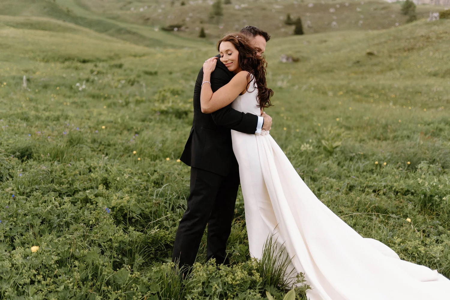 A bride and groom embracing outdoors in a green meadow, with rolling hills in the background, during a wedding celebration.