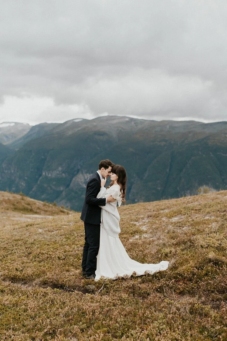 A couple dressed in wedding attire sharing an intimate moment outdoors in a mountainous landscape with overcast skies.