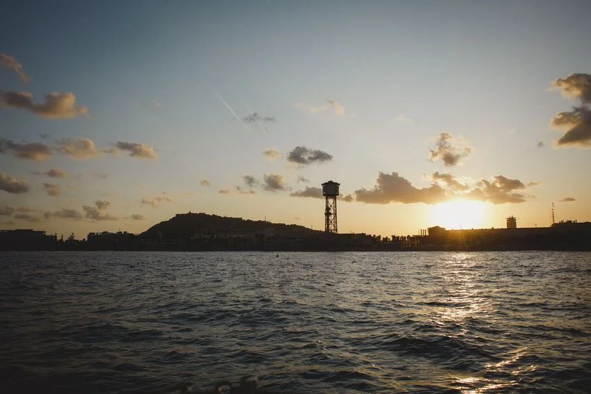 Sunset over water with a distant shoreline, a tower silhouette, and scattered clouds in the sky.