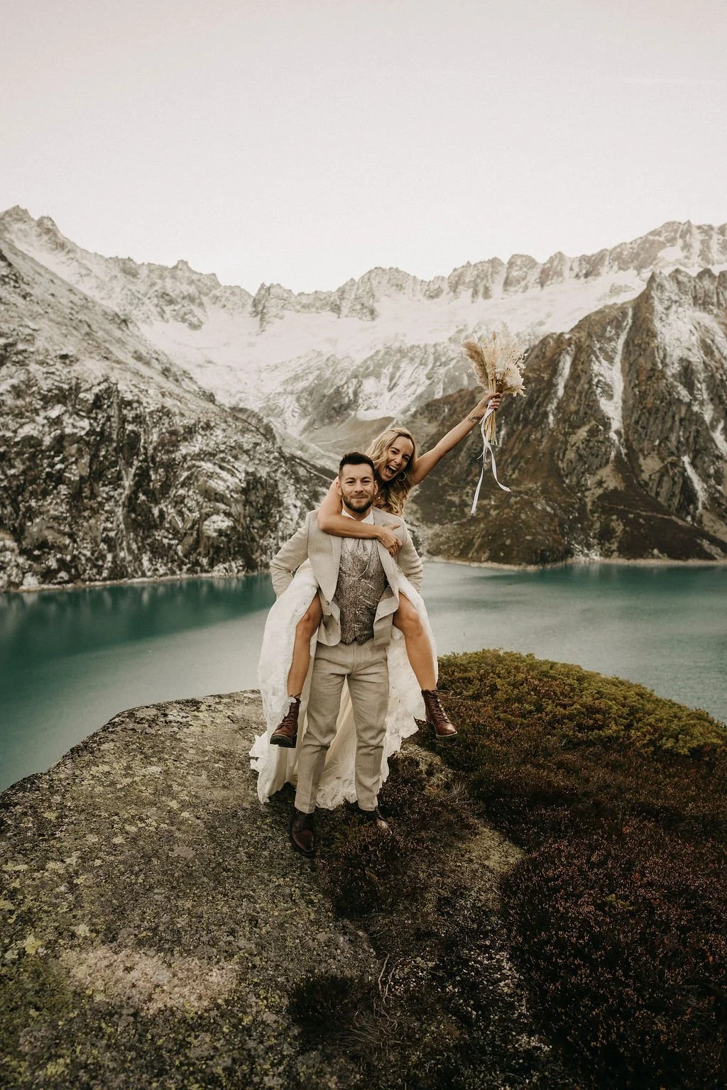 A couple in wedding attire celebrating outdoors on a rocky ledge near a lake with snow-capped mountains in the background; the woman is sitting on the man's shoulders, holding a bouquet, raising it joyfully.