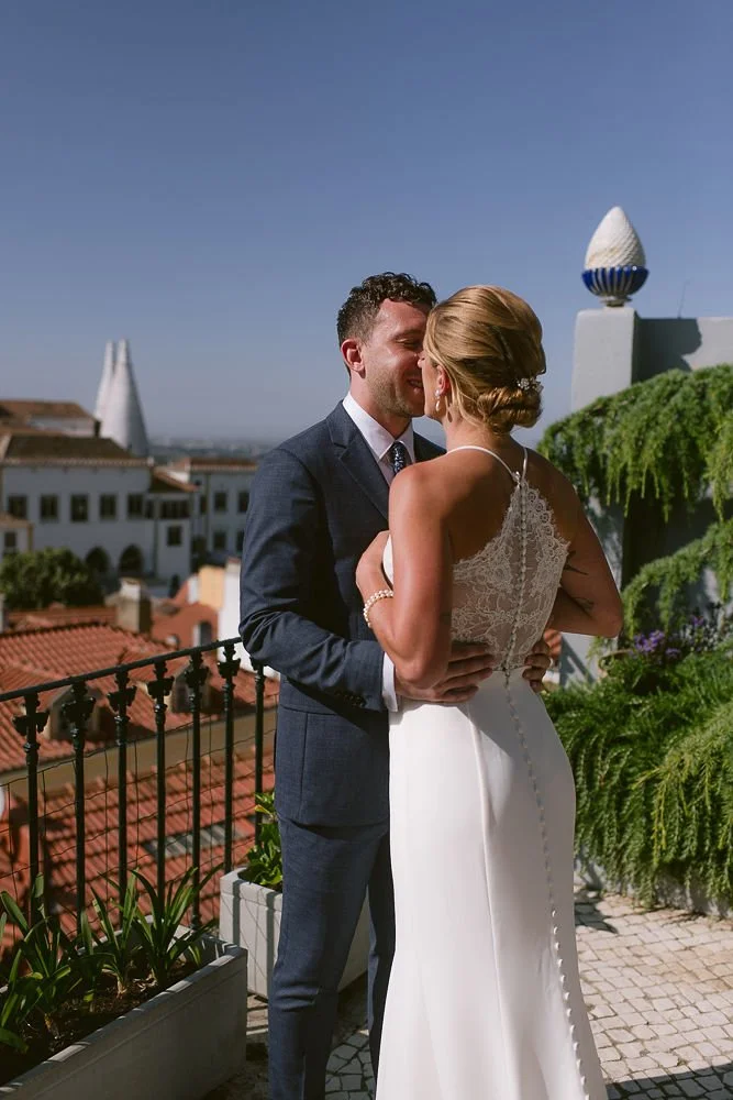 A bride and groom standing close together on a rooftop terrace, with the groom in a dark suit and the bride in a white wedding dress, embracing and whispering to each other during their wedding.