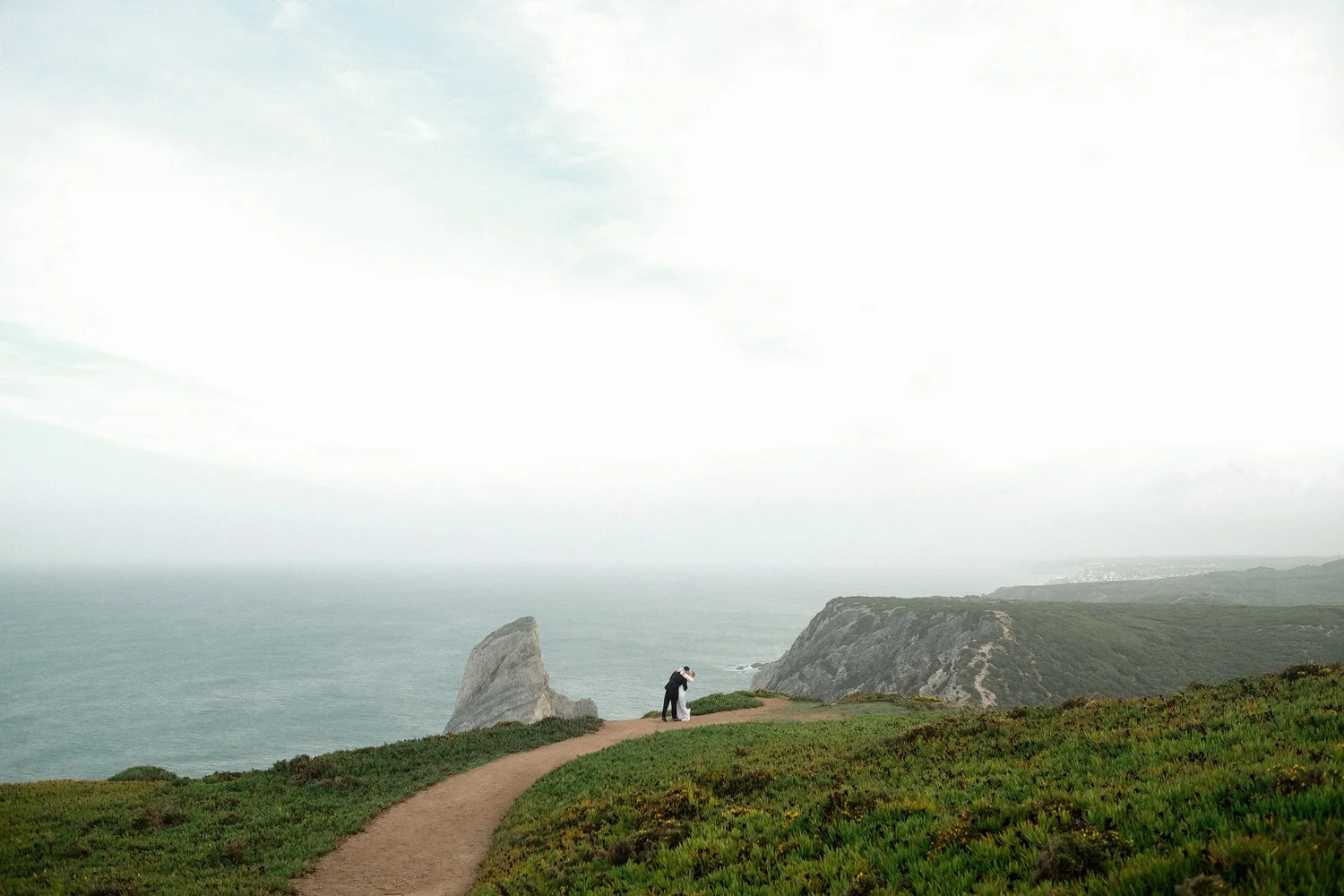 A couple hugging on a dirt path near cliffs overlooking the ocean, with a grassy area in the foreground and a cloudy sky overhead.
