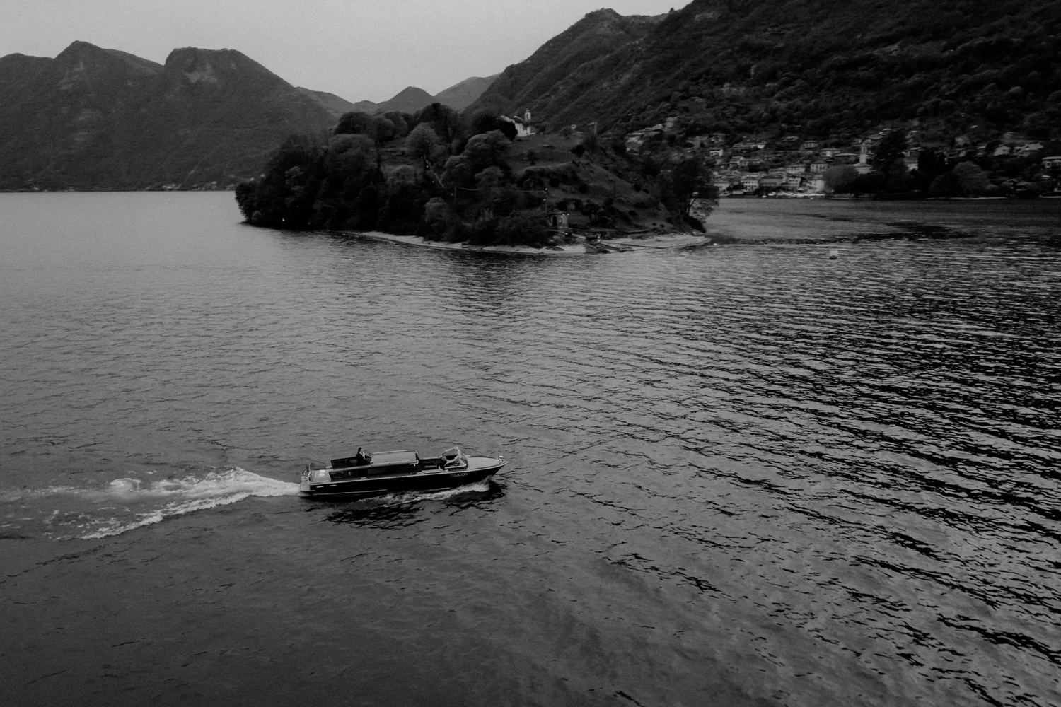 A boat moves across a body of water near a hilly shoreline with scattered buildings and trees.