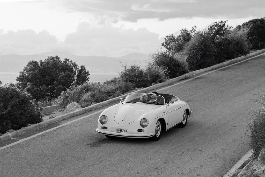 A vintage white convertible car driving on a winding road with two people inside, against a backdrop of trees and cloudy sky in black and white.