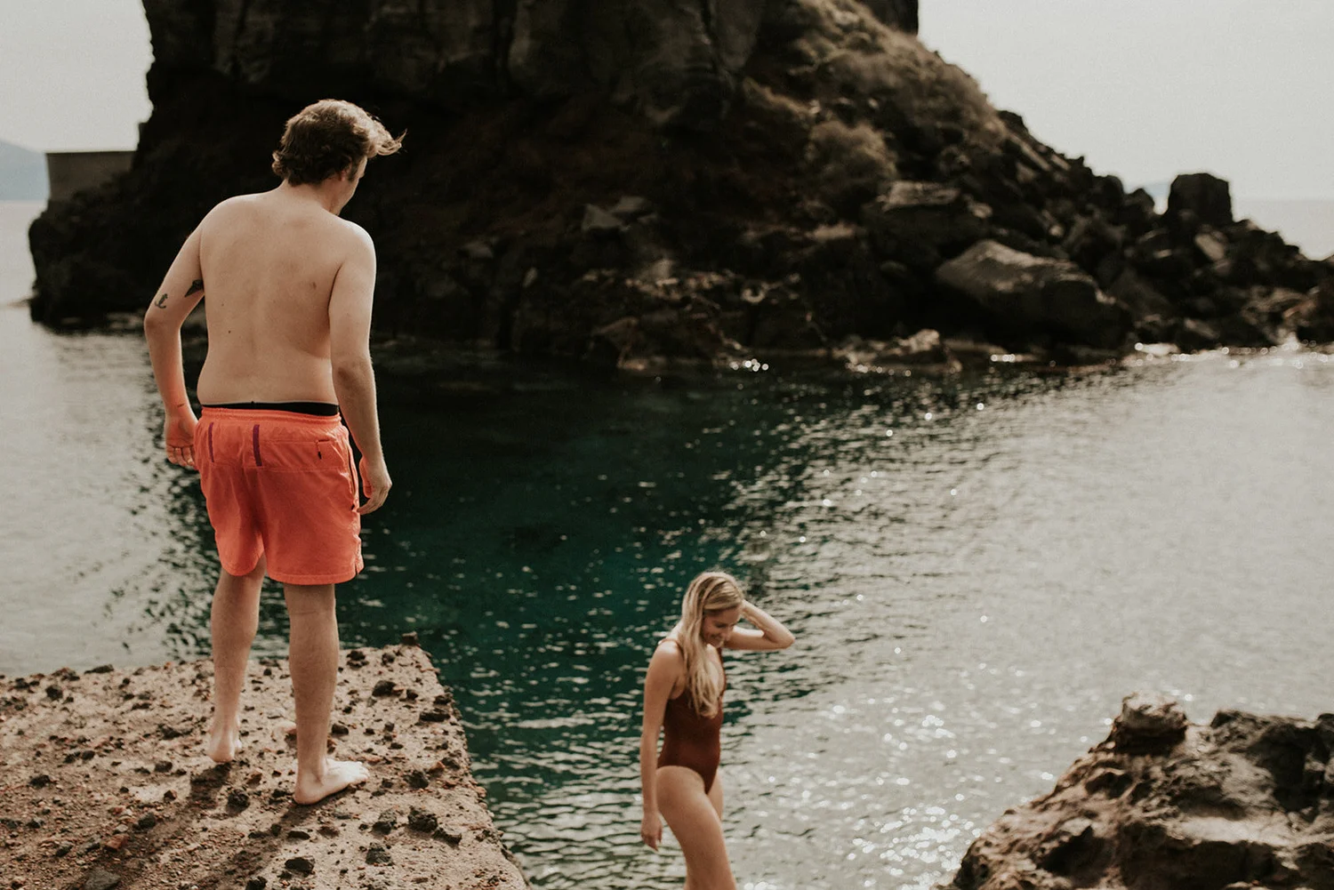 A man in orange shorts stands on a rocky cliff beside a woman in a swimsuit walking into the water near a rocky shoreline.
