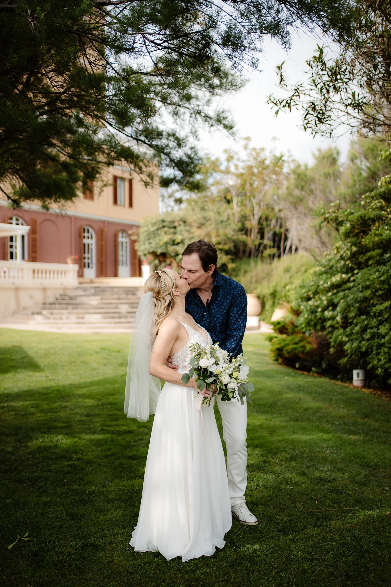 A bride and groom kiss outdoors on a green lawn, with trees and a pink building in the background.
