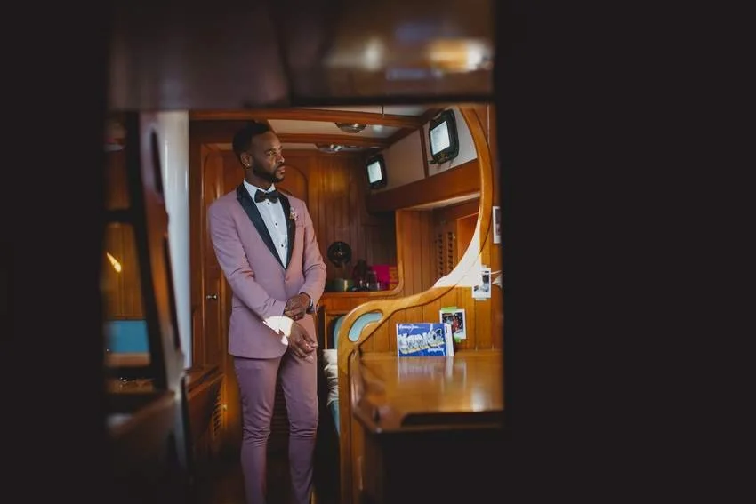 A man dressed in a pink suit with a black bow tie, standing inside a wooden-paneled room, possibly a yacht or boat, with a serious expression.