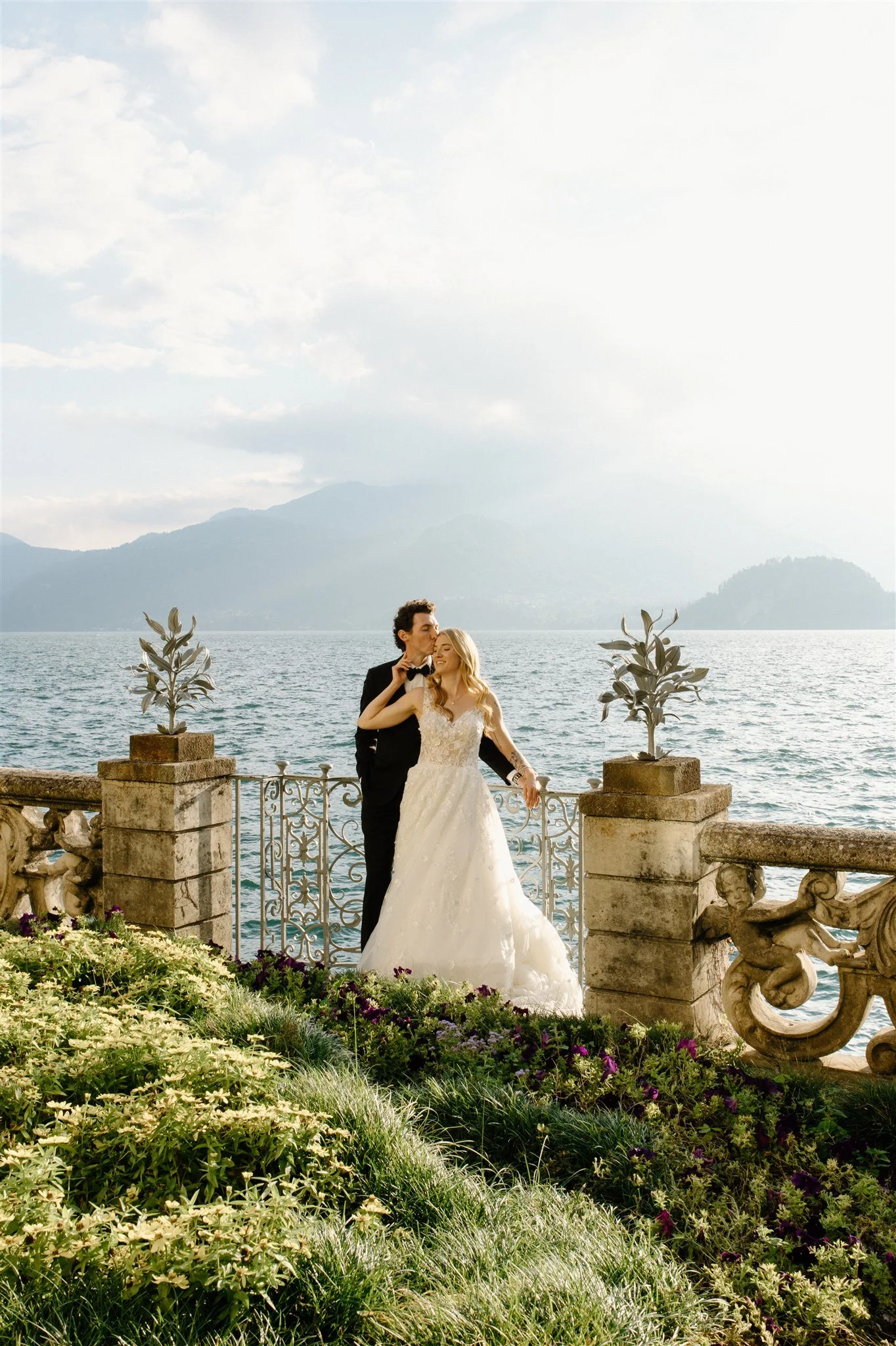 A newlywed couple stands on a decorative balcony by a lake with mountains in the background, surrounded by flowers and plants.