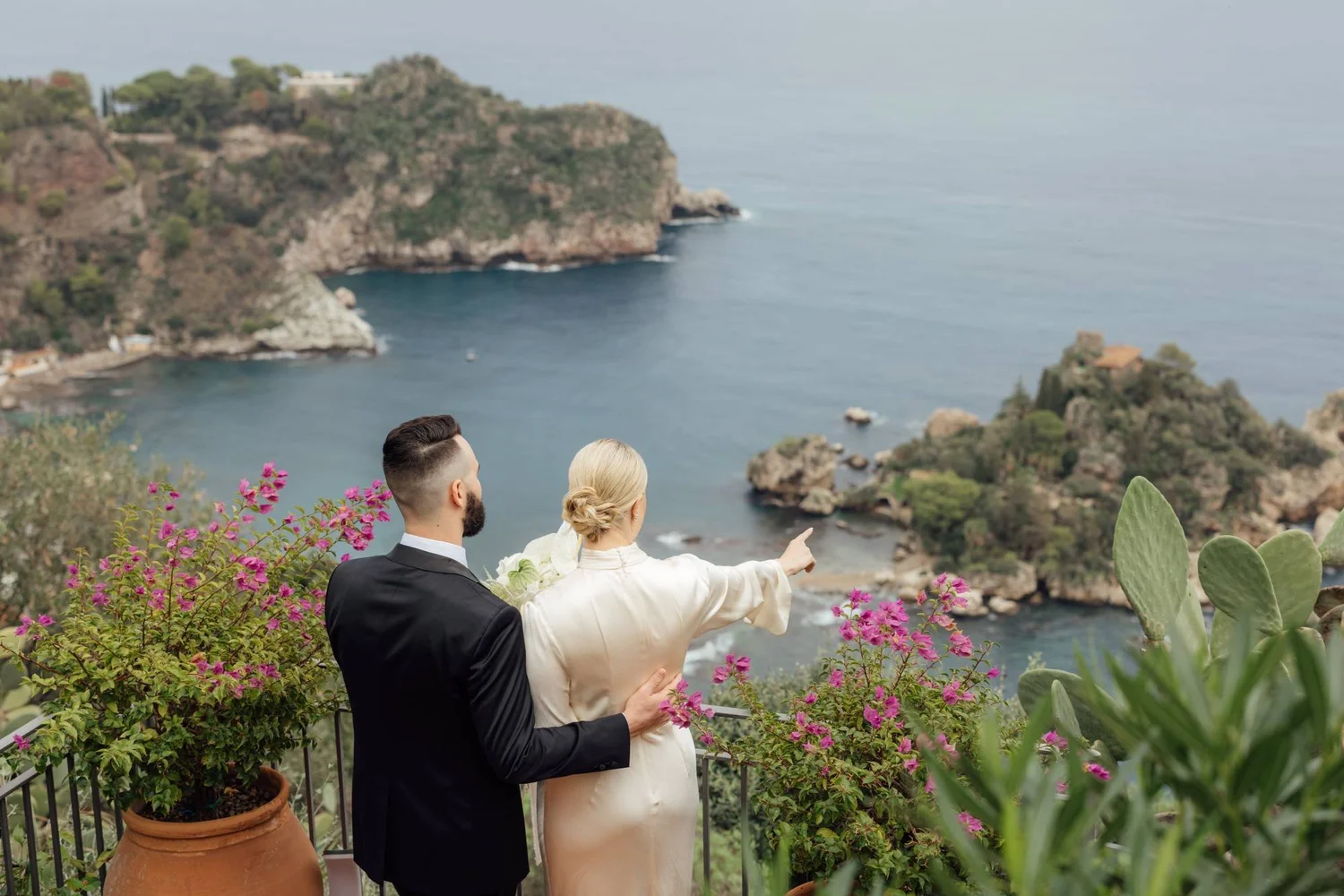 A couple, dressed formally, stands on a terrace with pink flowers and green cactus, overlooking a coastal area with cliffs, water, and small houses, during daytime.