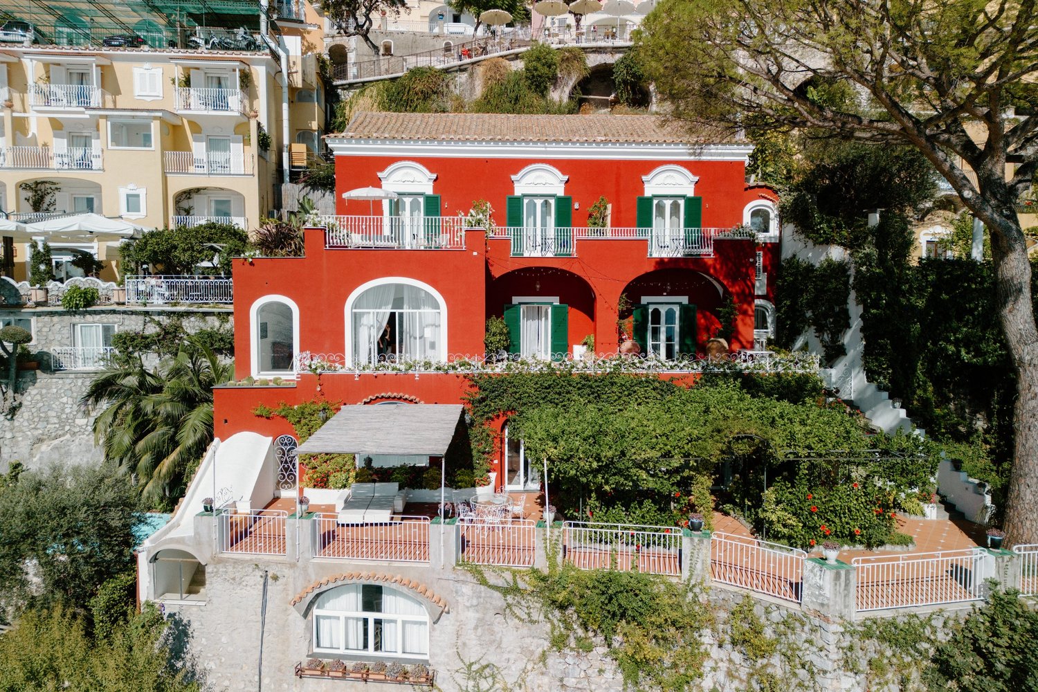 A colorful hillside building with a red facade, green window shutters, and several balconies with white railings, set among lush greenery and large trees.