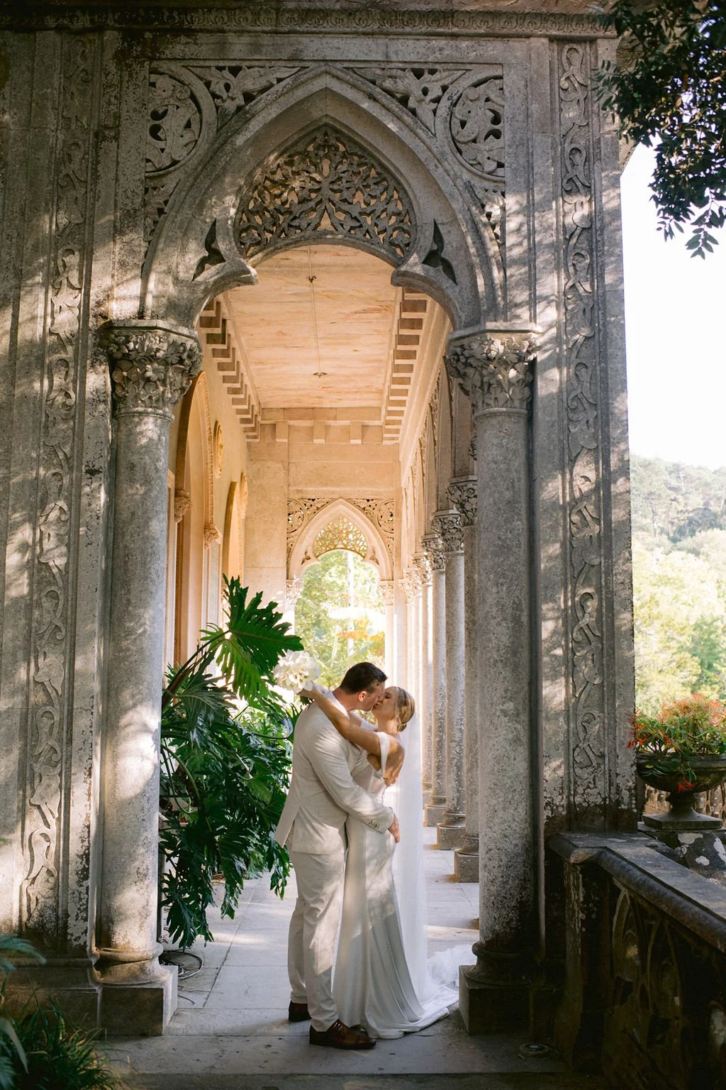 A wedding couple in white attire sharing a kiss under an ornate stone archway with intricate carvings, surrounded by lush greenery and soft sunlight.