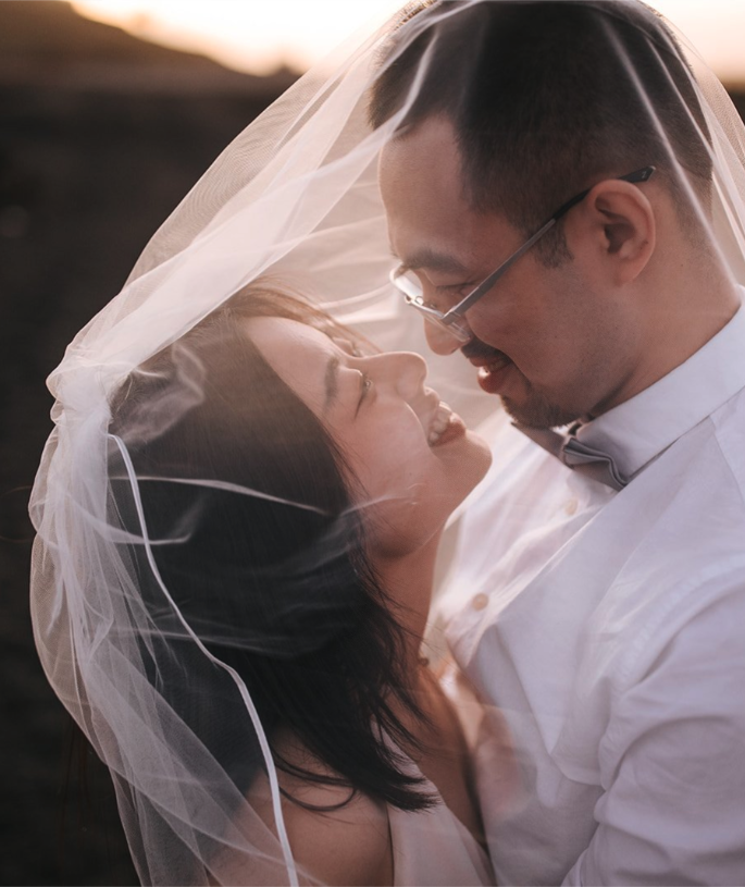A couple smiling and looking into each other's eyes under a wedding veil, outdoors during sunset.
