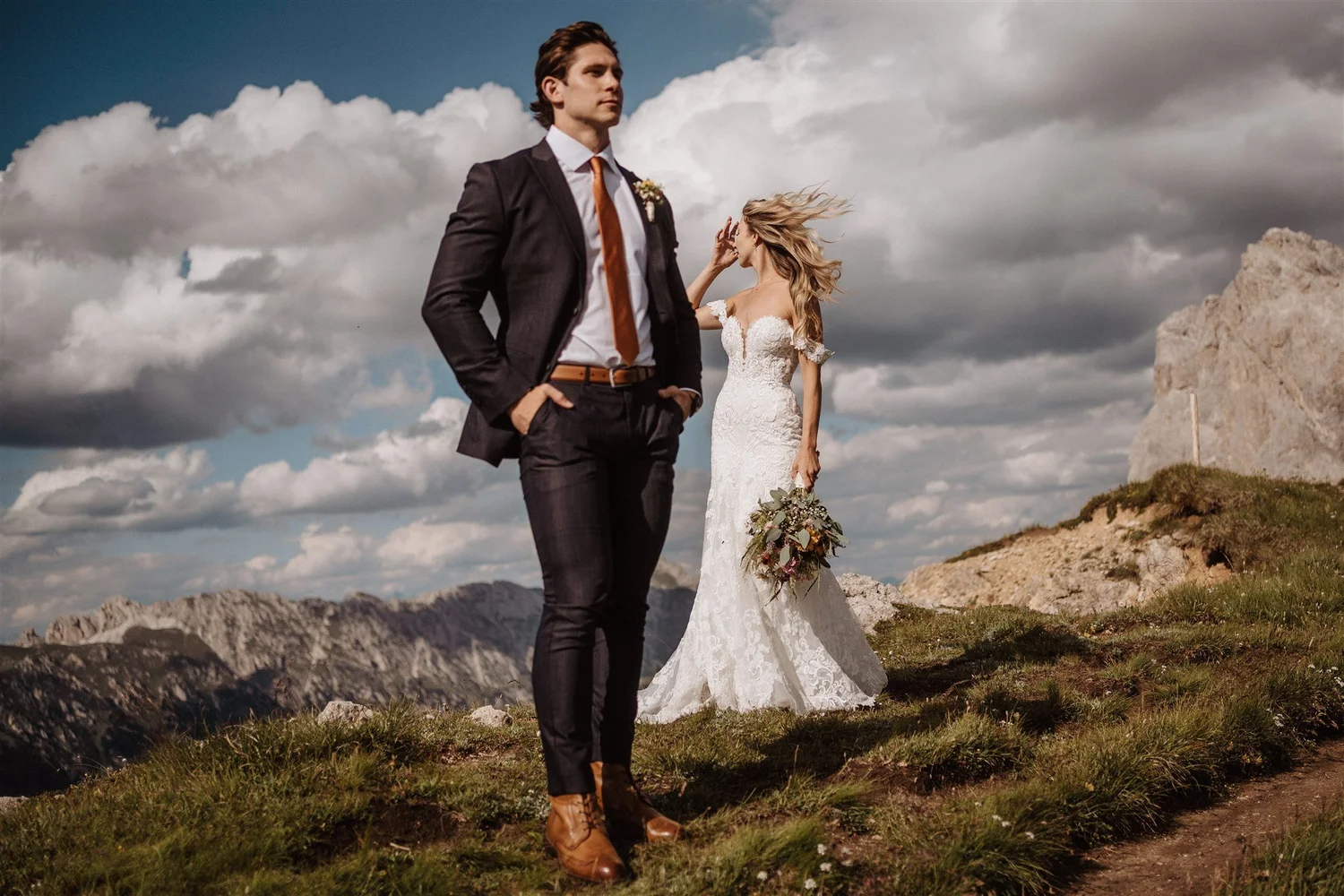 A man in a suit with hands in pockets and a woman in a wedding dress holding a bouquet, standing outdoors on a grassy area with mountains and cloudy sky in the background.