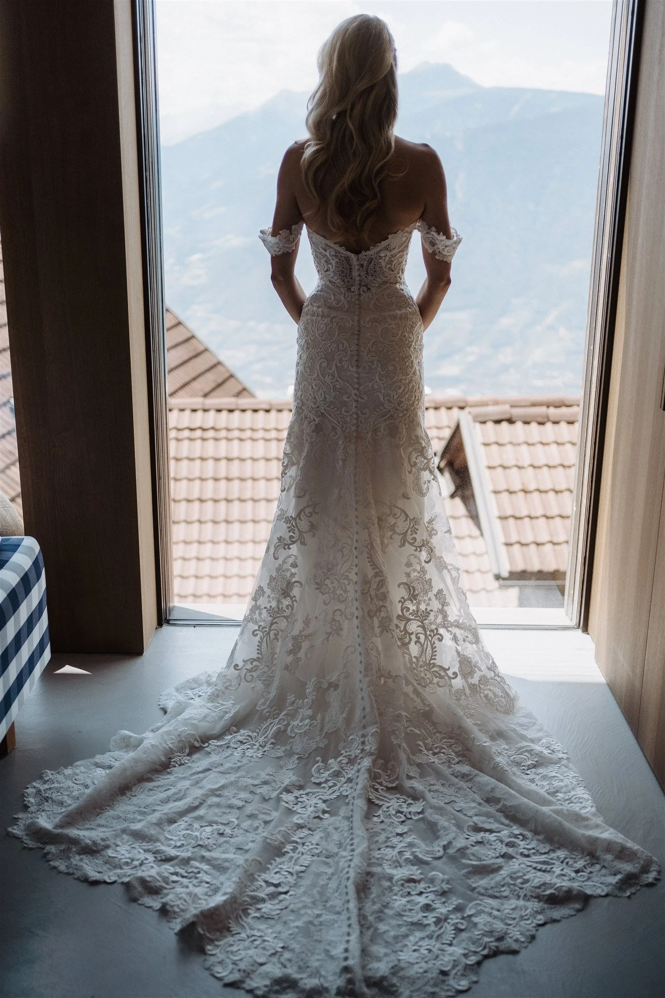 A bride in a lace wedding dress standing at a window, looking out at distant mountains.