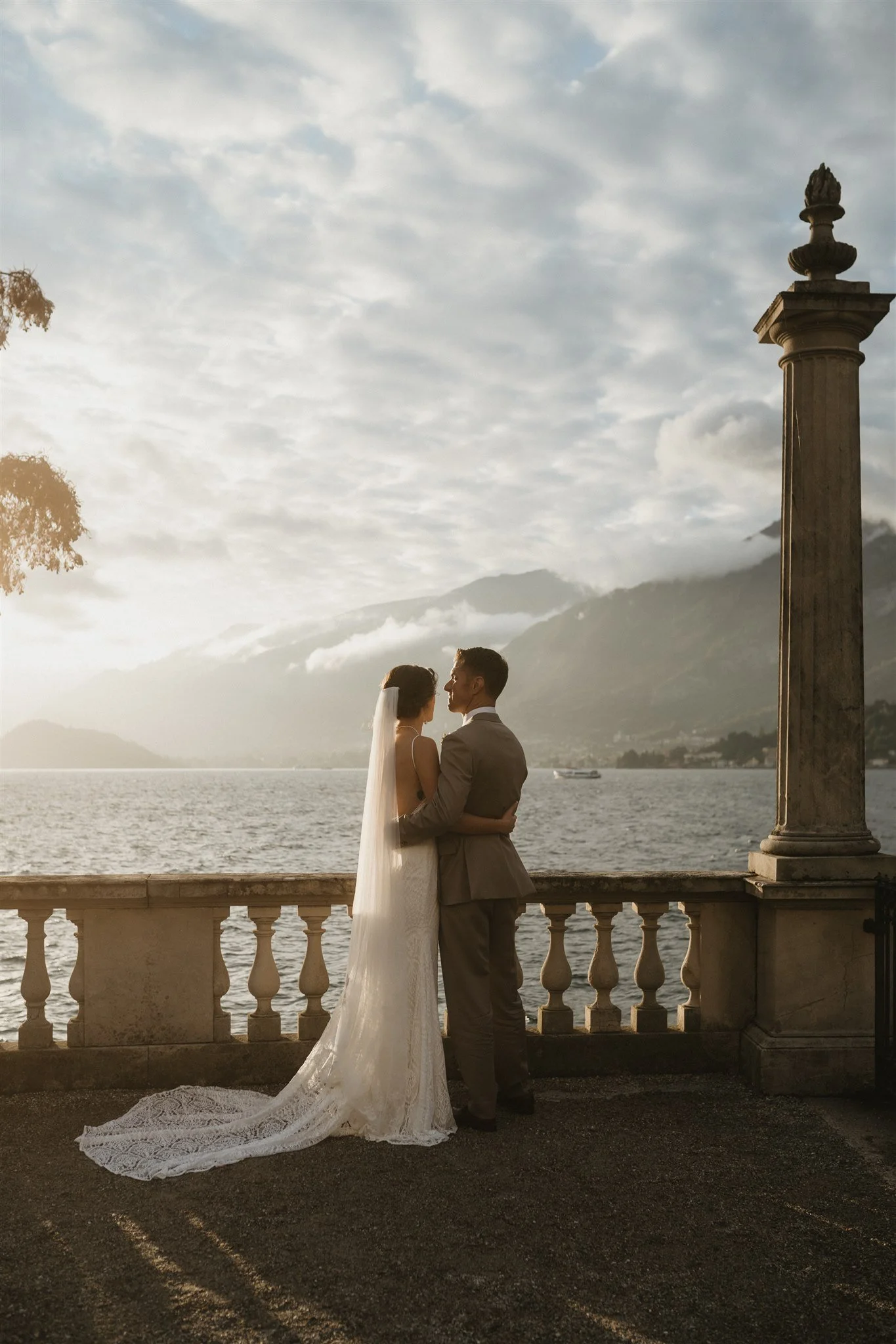 A bride and groom stand close together on a balcony overlooking a body of water, with mountains in the distance, during sunset, with cloudy sky above.