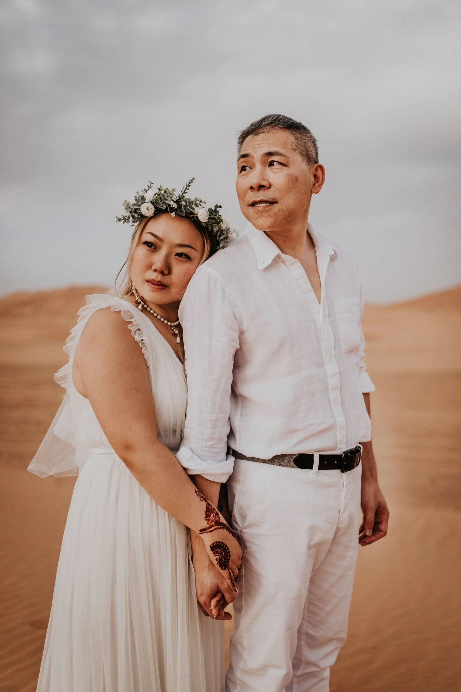 A couple dressed in white standing close together outdoors in a desert landscape with sand dunes in the background. The woman is wearing a white dress with ruffled sleeves, a floral crown, and pearl jewelry, and has henna tattoos on her hand and wris