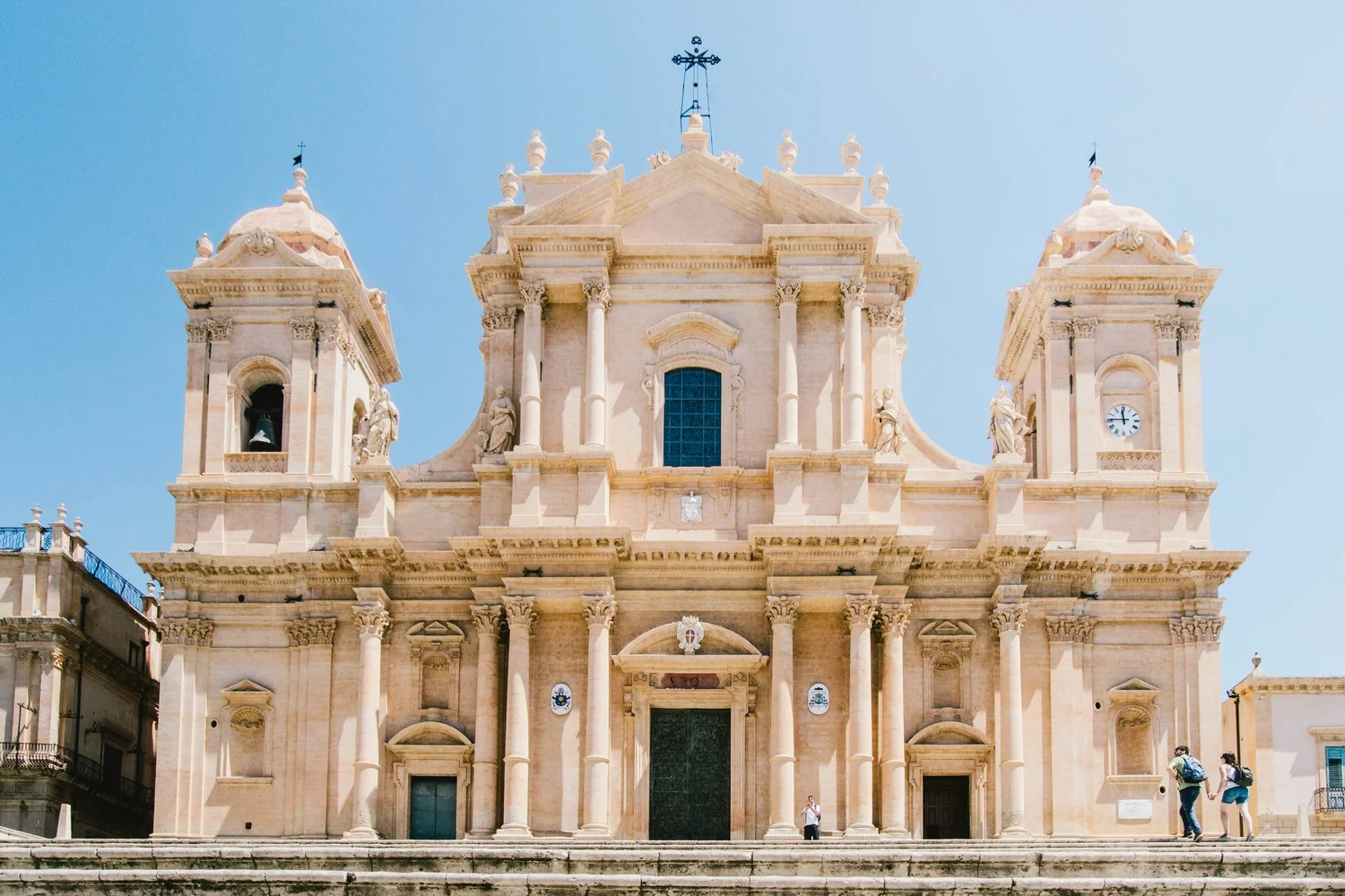 Facade of a historic church with ornate architectural details, statues, columns, and a clock, set against a clear blue sky with a few visitors at the entrance.