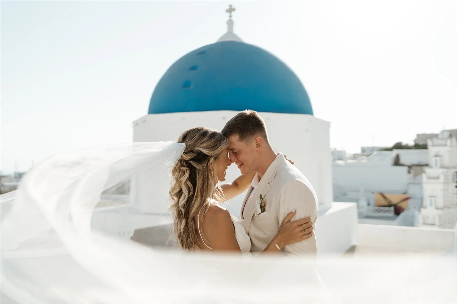 A couple dressed in wedding attire sharing an intimate moment on a white rooftop with a blue-domed building in the background.