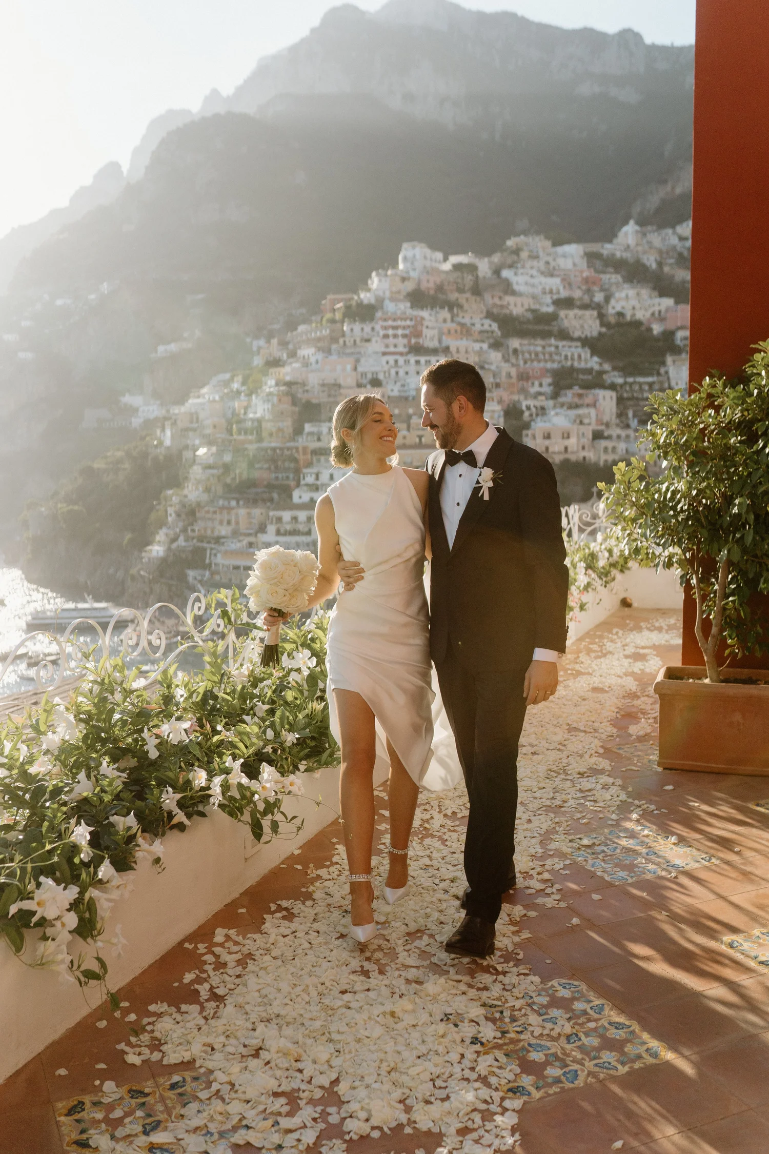 A newlywed couple walking on a flower petal-strewn outdoor terrace with a scenic hillside town in the background, during sunset.
