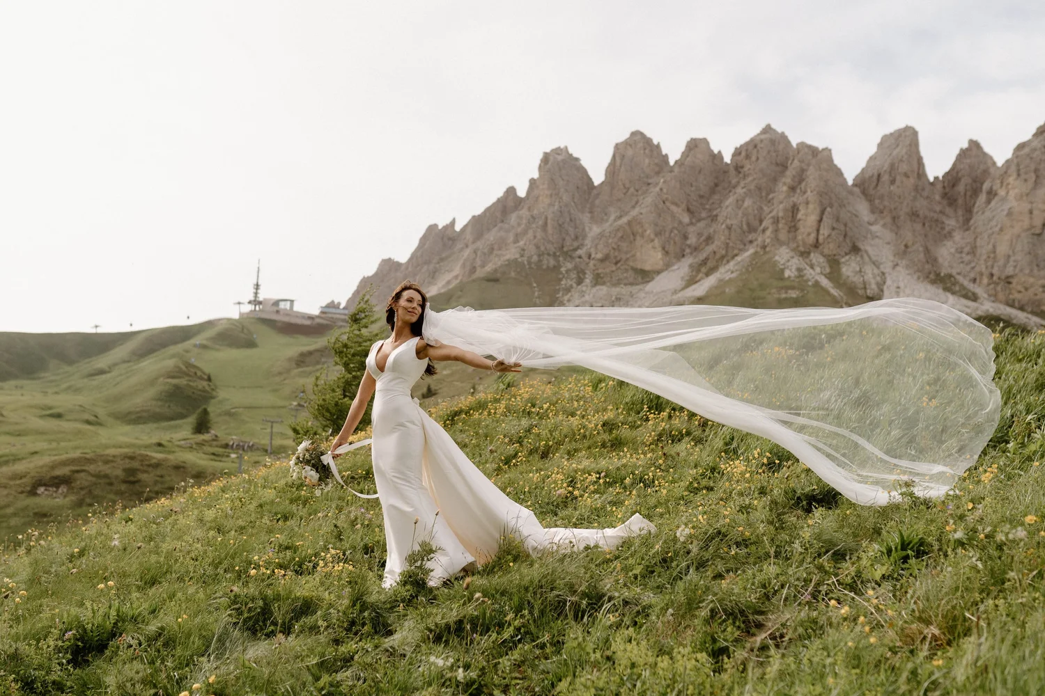 A bride in a white wedding dress standing in a grassy field with yellow flowers, holding a bouquet, and wearing a long veil flowing in the wind, with mountain peaks in the background under an overcast sky.