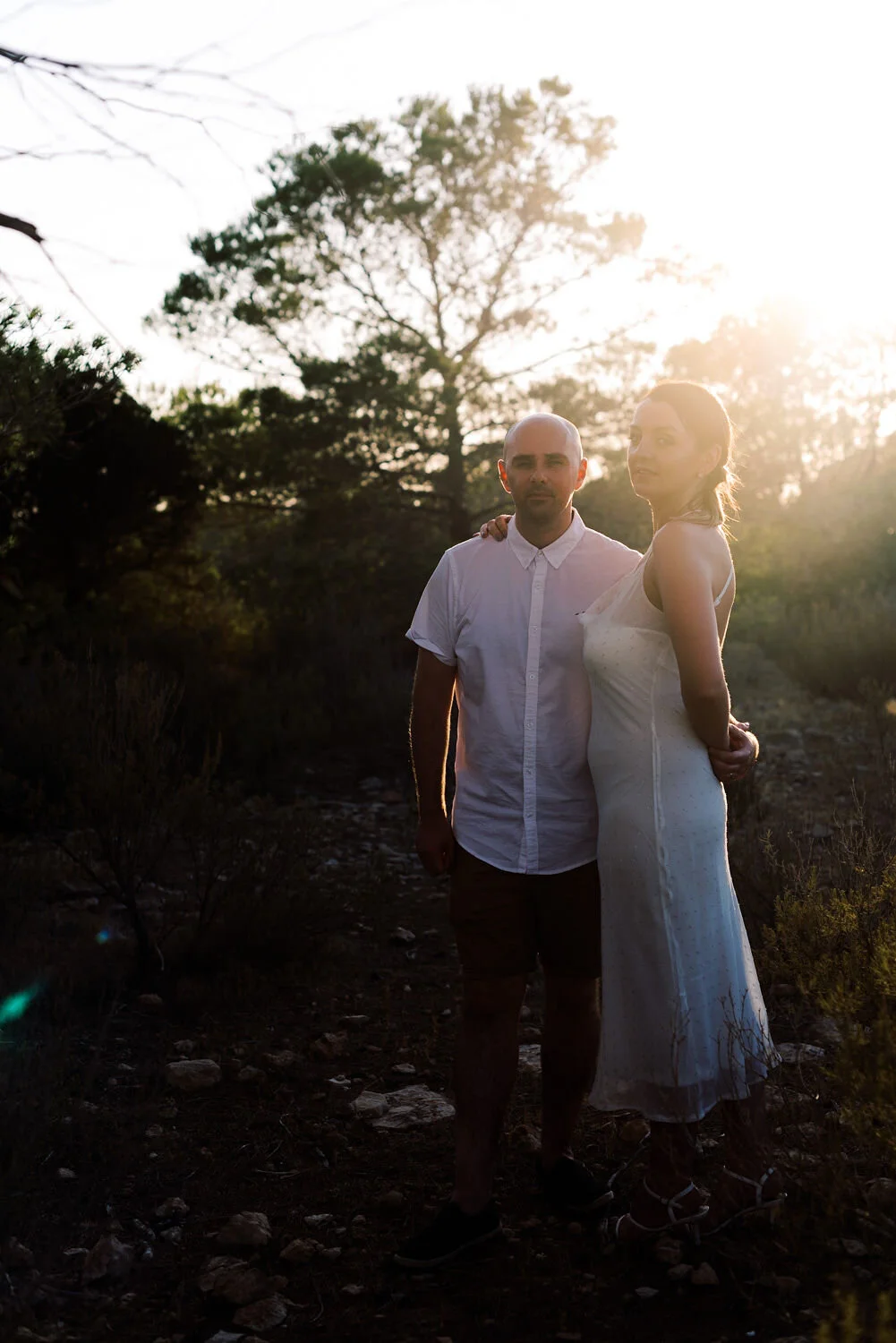 A man and woman standing close together outdoors during sunset, surrounded by trees and bushes. The woman is wearing a white dress, and the man is dressed in a white shirt and dark shorts. The sunlight creates a backlit effect, casting a warm glow ar