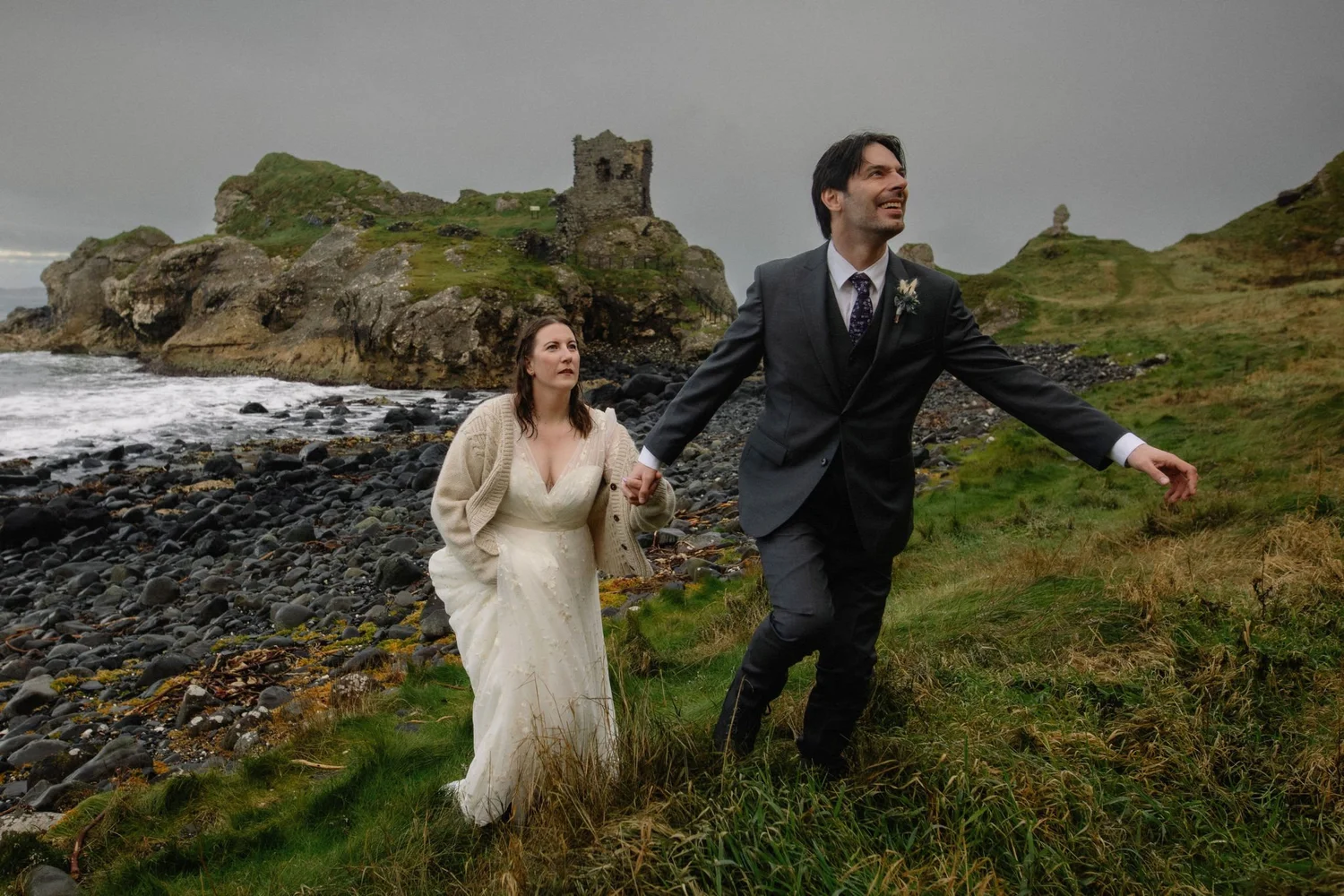 A couple dressed in wedding attire holding hands and walking along a rocky beach with green hills and ruins in the background.
