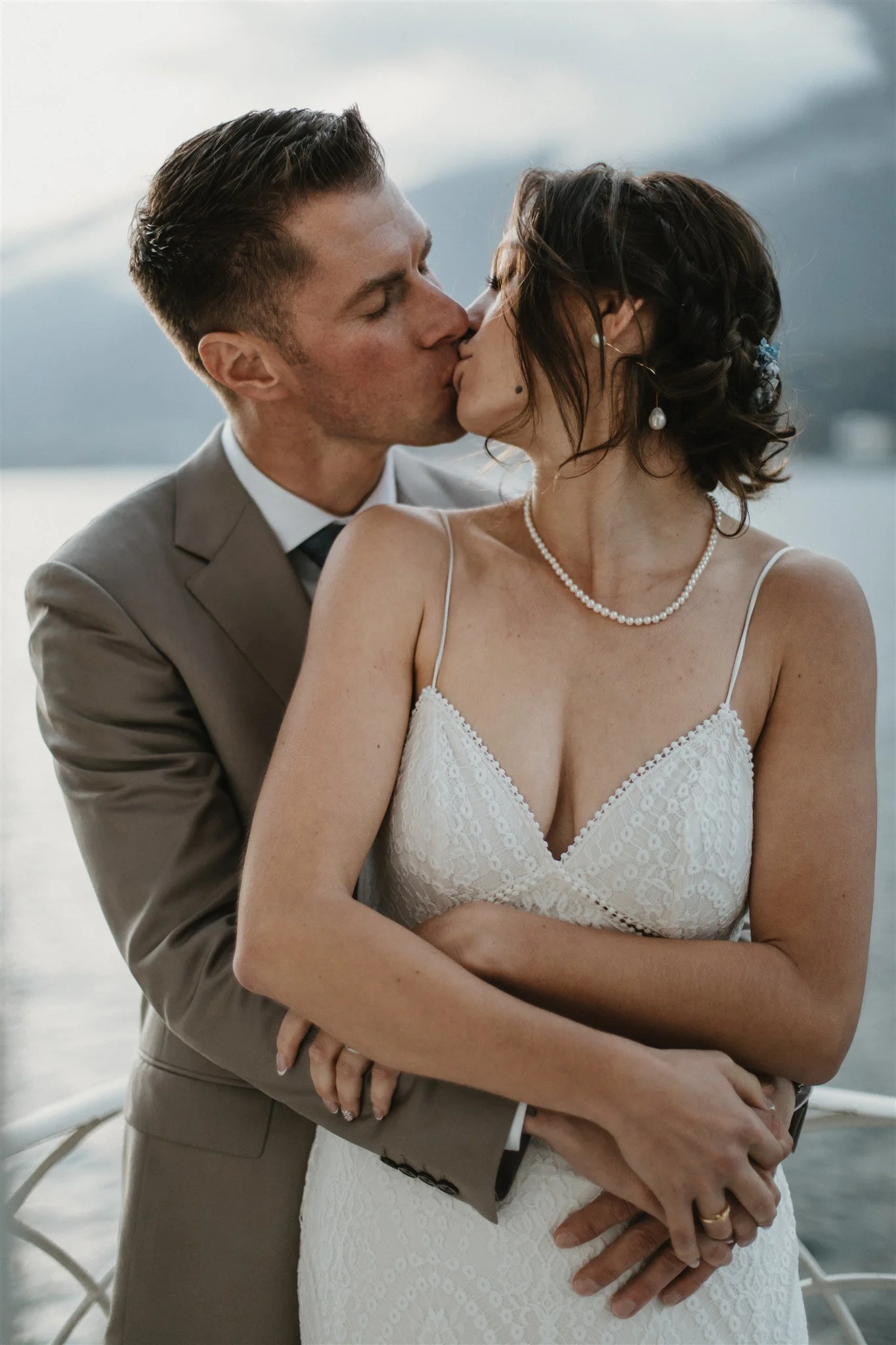 A newlywed couple sharing a kiss, with the groom embracing the bride from behind. The groom is in a light gray suit with a white shirt and dark tie, and the bride is in a white lace wedding dress with spaghetti straps, wearing pearl earrings and a pe