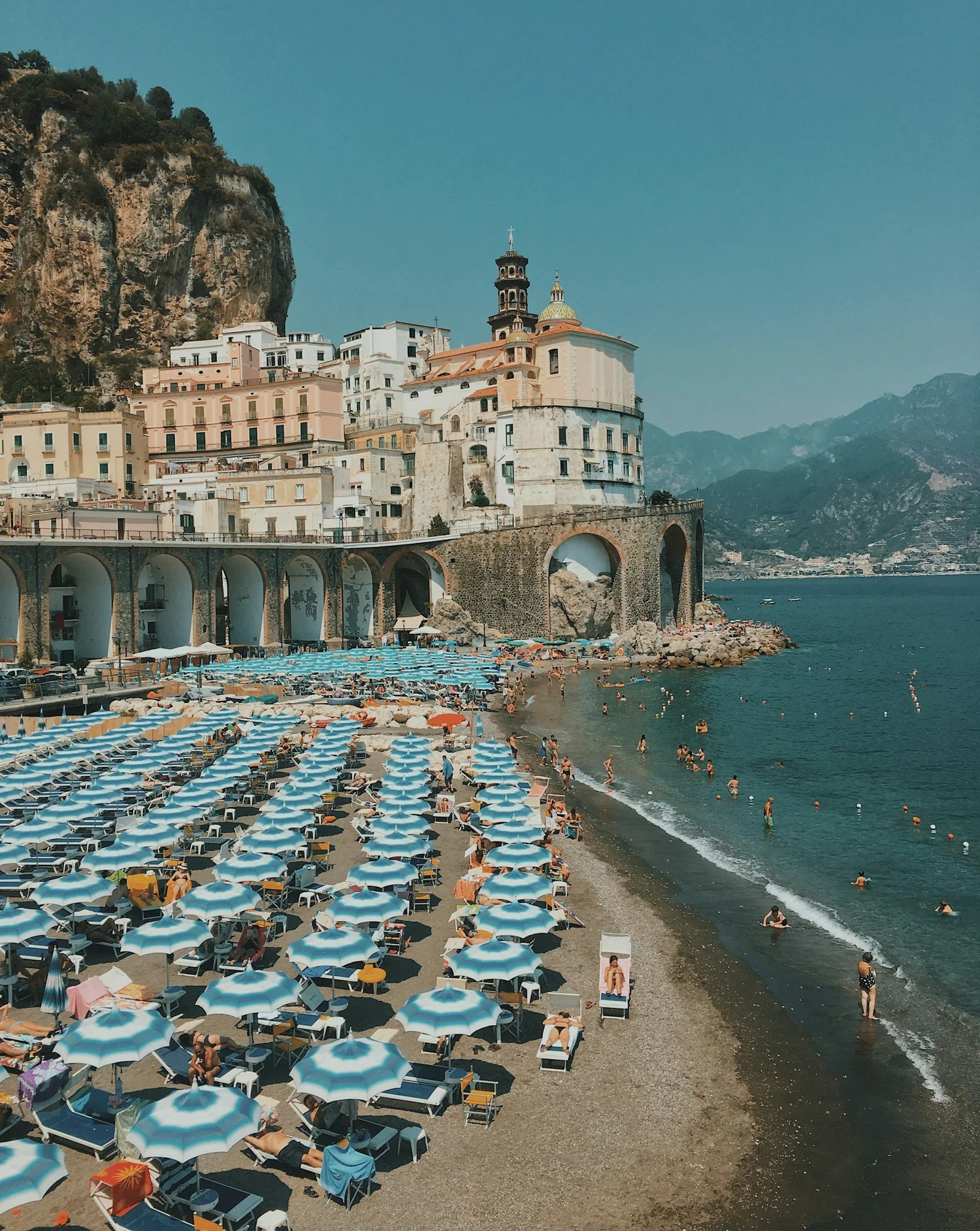 Beach with numerous blue and white striped umbrellas and people swimming and relaxing near the shoreline. Historical buildings and a rocky hillside in the background under a clear blue sky.