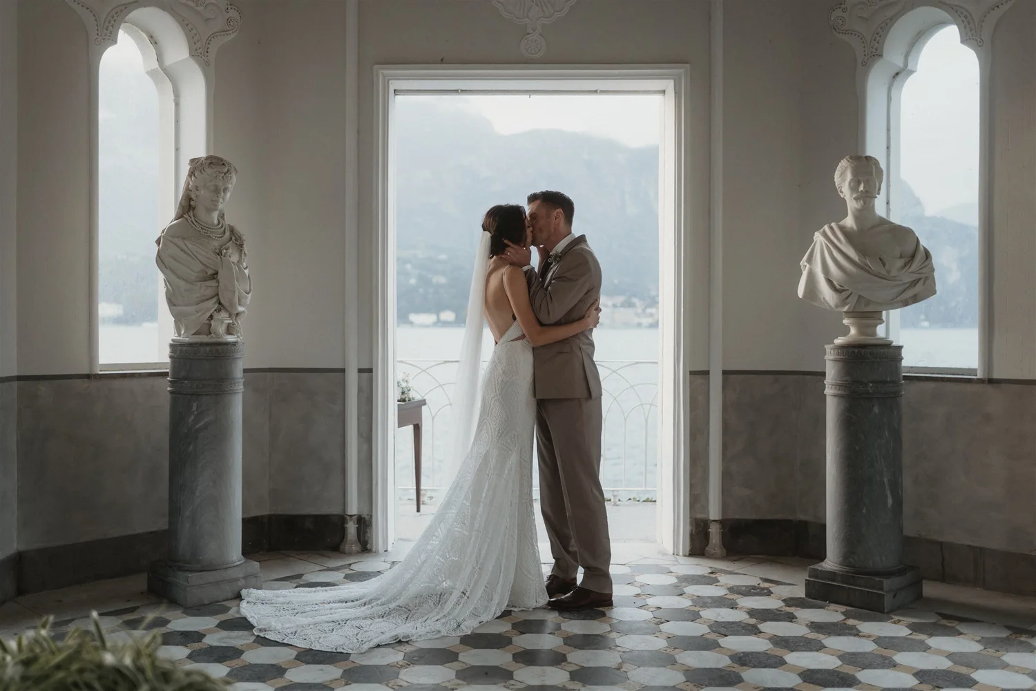 A bride and groom kiss inside a building with large windows and classical sculptures on pedestals.