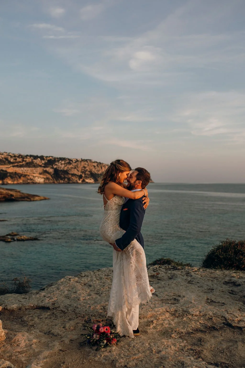 A couple on their wedding day, with the bride being carried by the groom, standing on a rocky ledge overlooking the ocean at sunset.