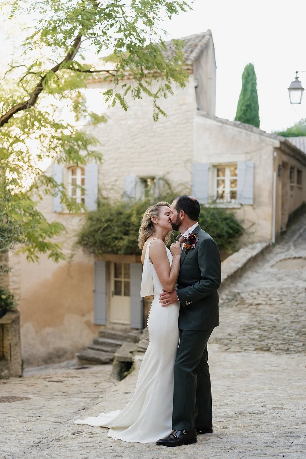 A bride and groom kiss in front of an old stone building with shutters on the windows, surrounded by greenery and a cobblestone street, during a wedding celebration.