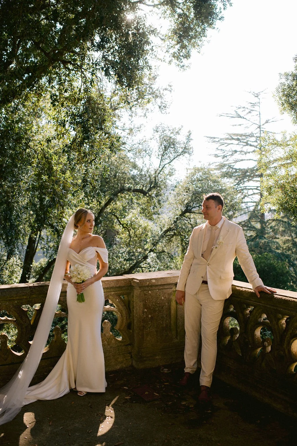 A bride and groom in wedding attire standing on a stone balcony surrounded by trees, with sunlight filtering through the leaves.