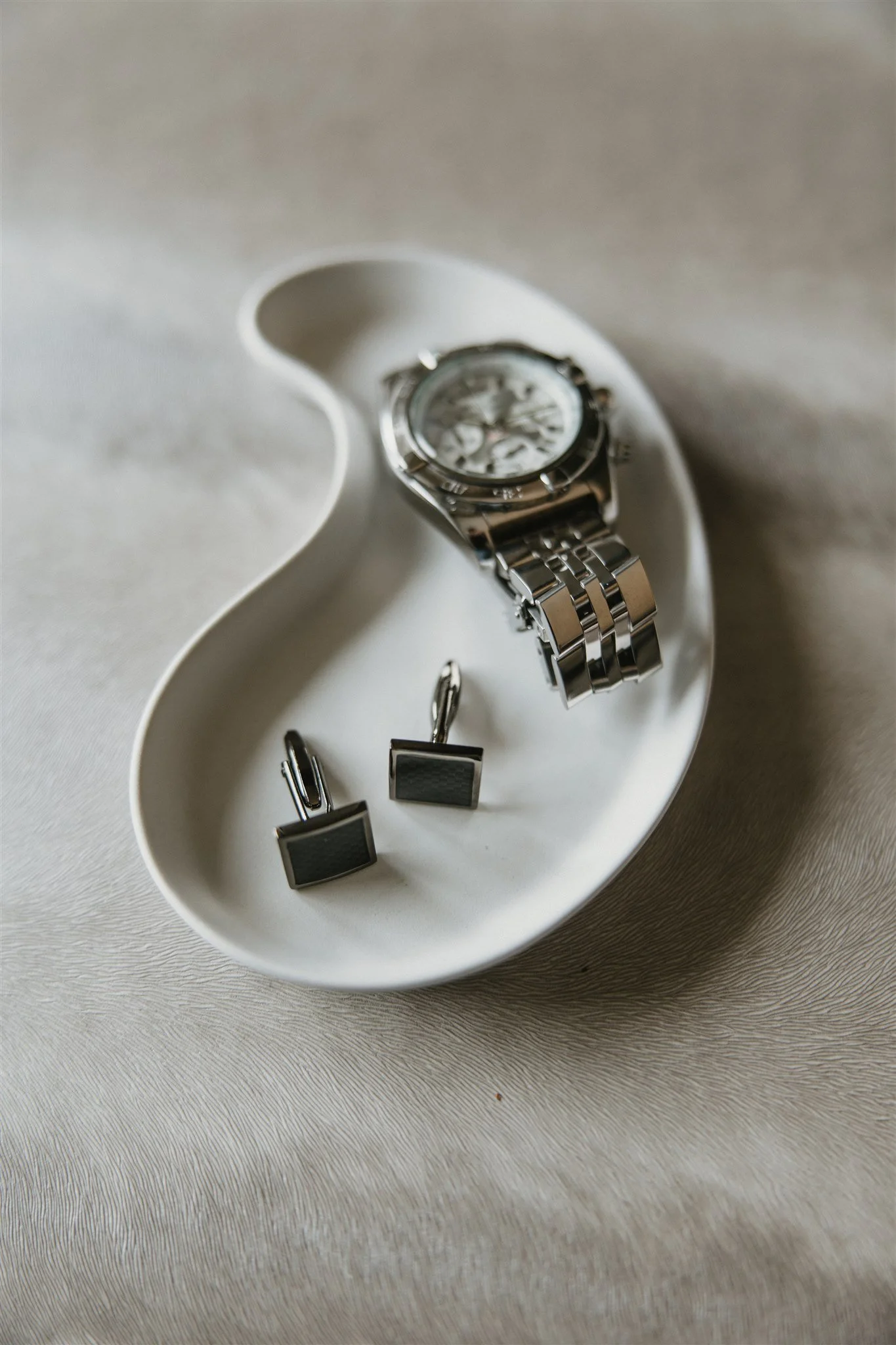 A display tray holding a silver wristwatch, a pair of black and silver cufflinks, and a pair of black and silver earrings, on a light-colored textured surface.