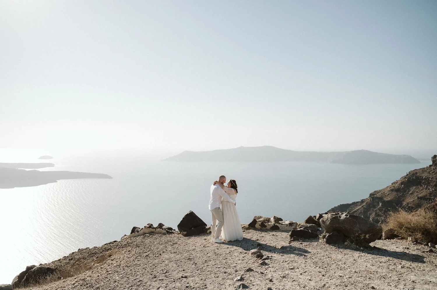 A couple dressed in white embracing on rocky terrain with a scenic water and mountainous landscape in the background.