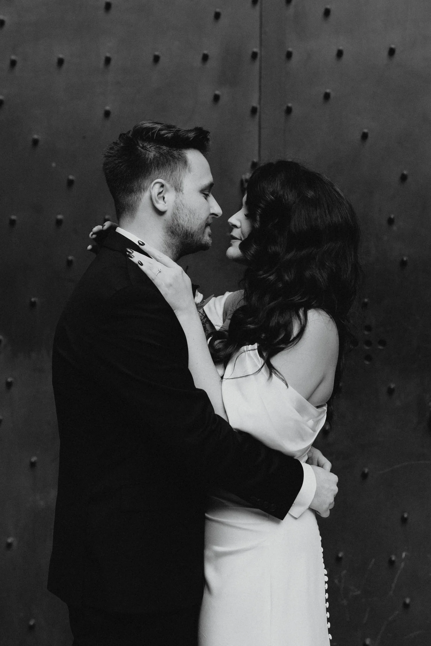 A black and white photograph of a couple holding each other intimately. The man has short hair and a beard, dressed in a suit, and the woman has long, dark, wavy hair, wearing a strapless dress. They are standing close to each other, with their faces almost touching, against a dark textured background.