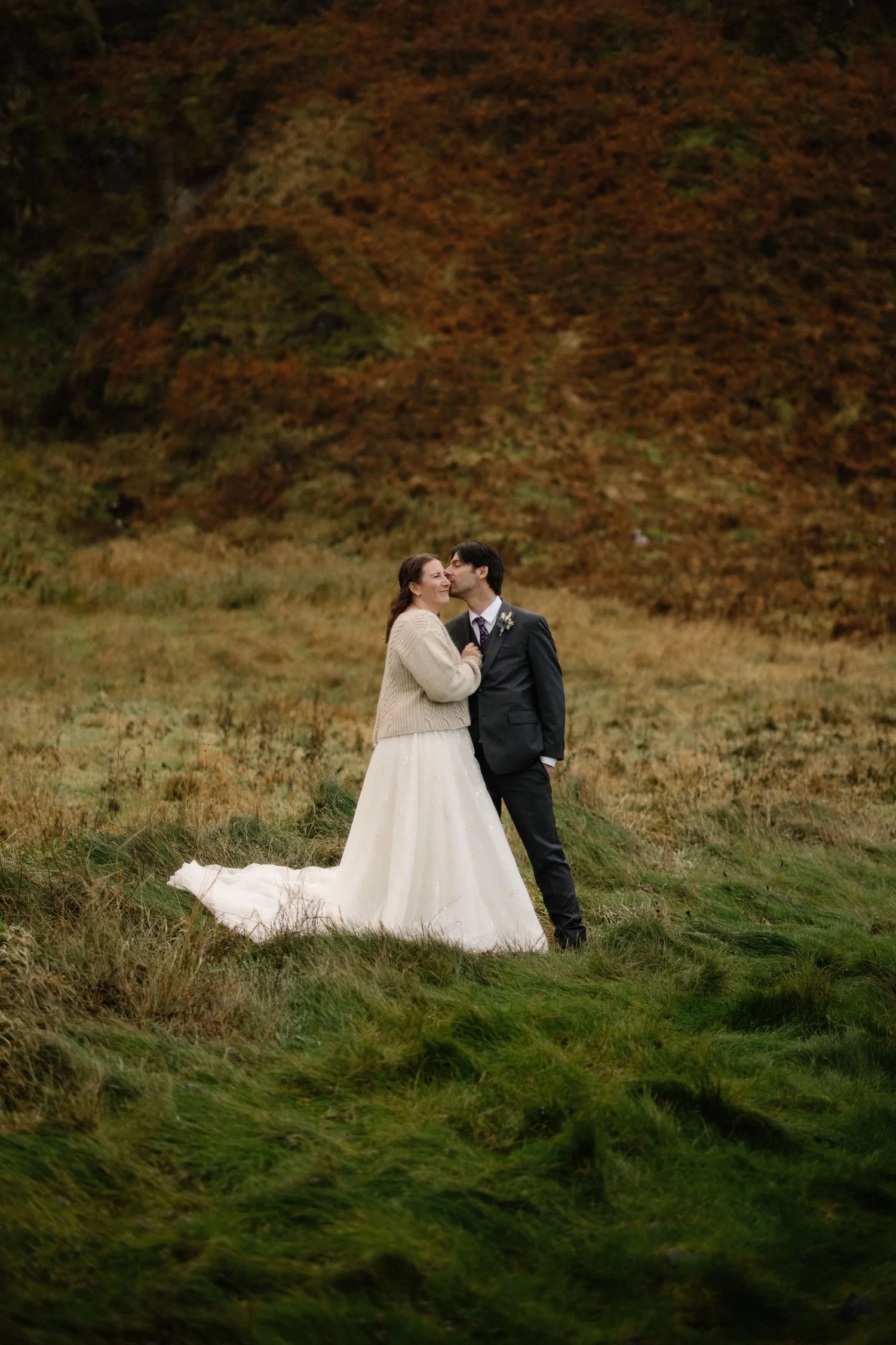 A couple in wedding attire standing in a grassy field surrounded by fall-colored trees, with the man giving the woman a gentle kiss on the cheek.