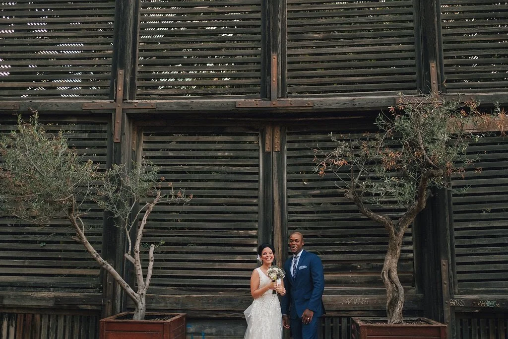 A couple in wedding attire standing outdoors in front of a wooden structure with two trees in planters.