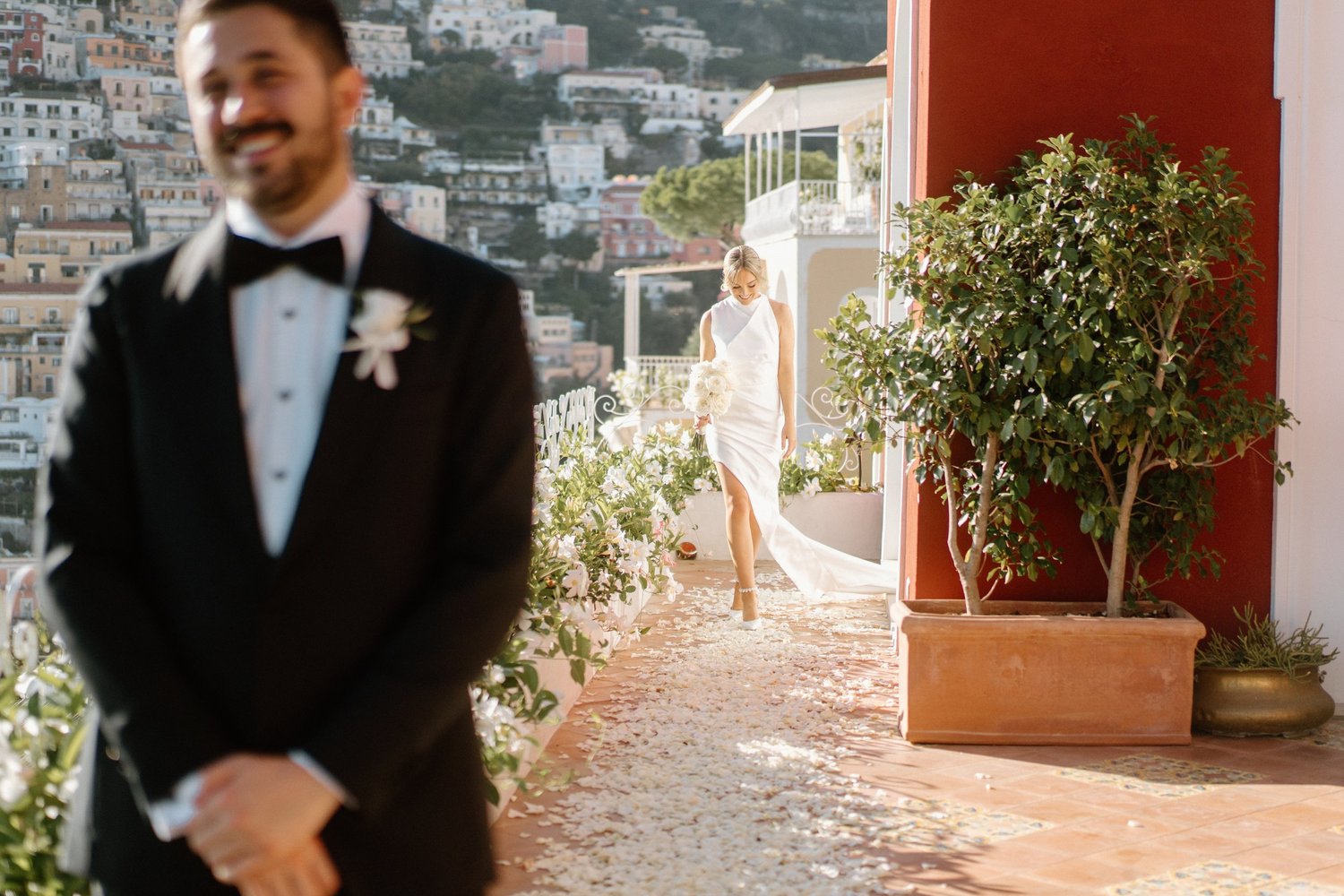 A bride in a white wedding dress holding a bouquet walking towards a man in a tuxedo, with a cityscape background and a potted plant on the right.