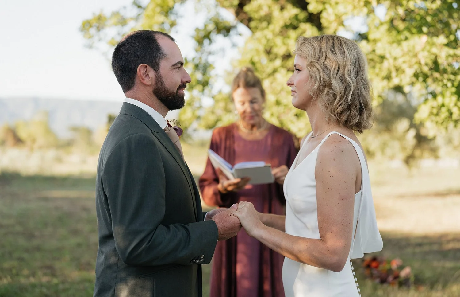 A couple getting married outdoors, holding hands and smiling at each other as a officiant officiates in the background.