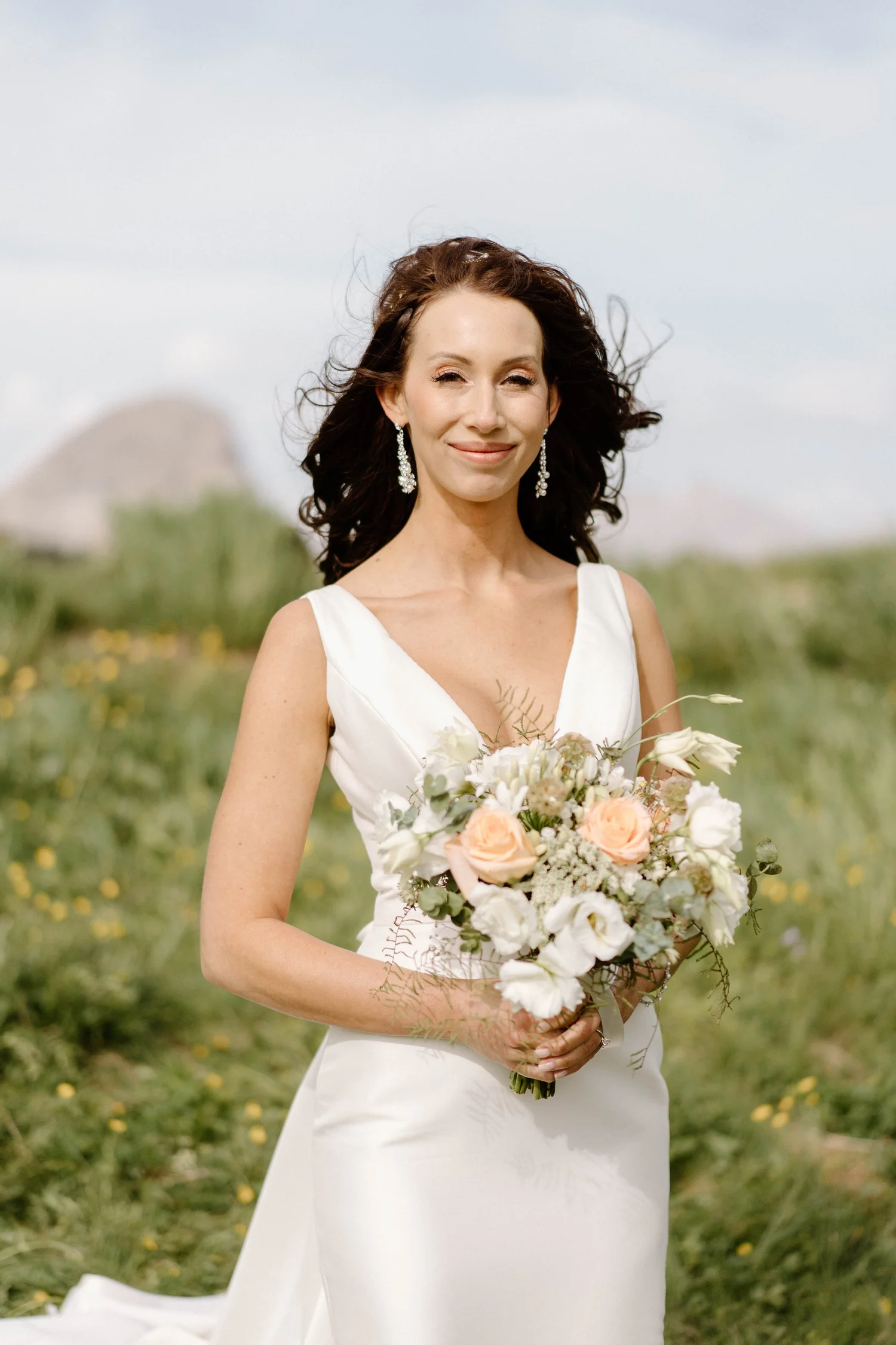 A smiling woman in a white wedding dress holding a bouquet of flowers outdoors on a grassy field with hills in the background.