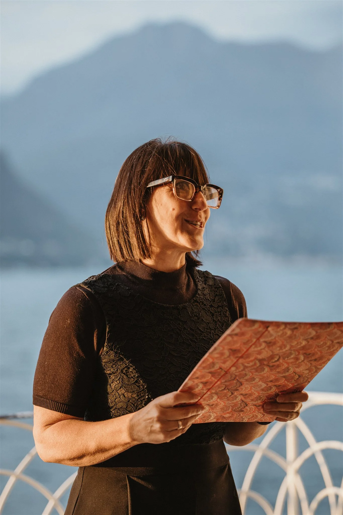 A woman wearing glasses and a black lace top reading a patterned menu near a waterfront with wind turbines and a blue sky in the background.