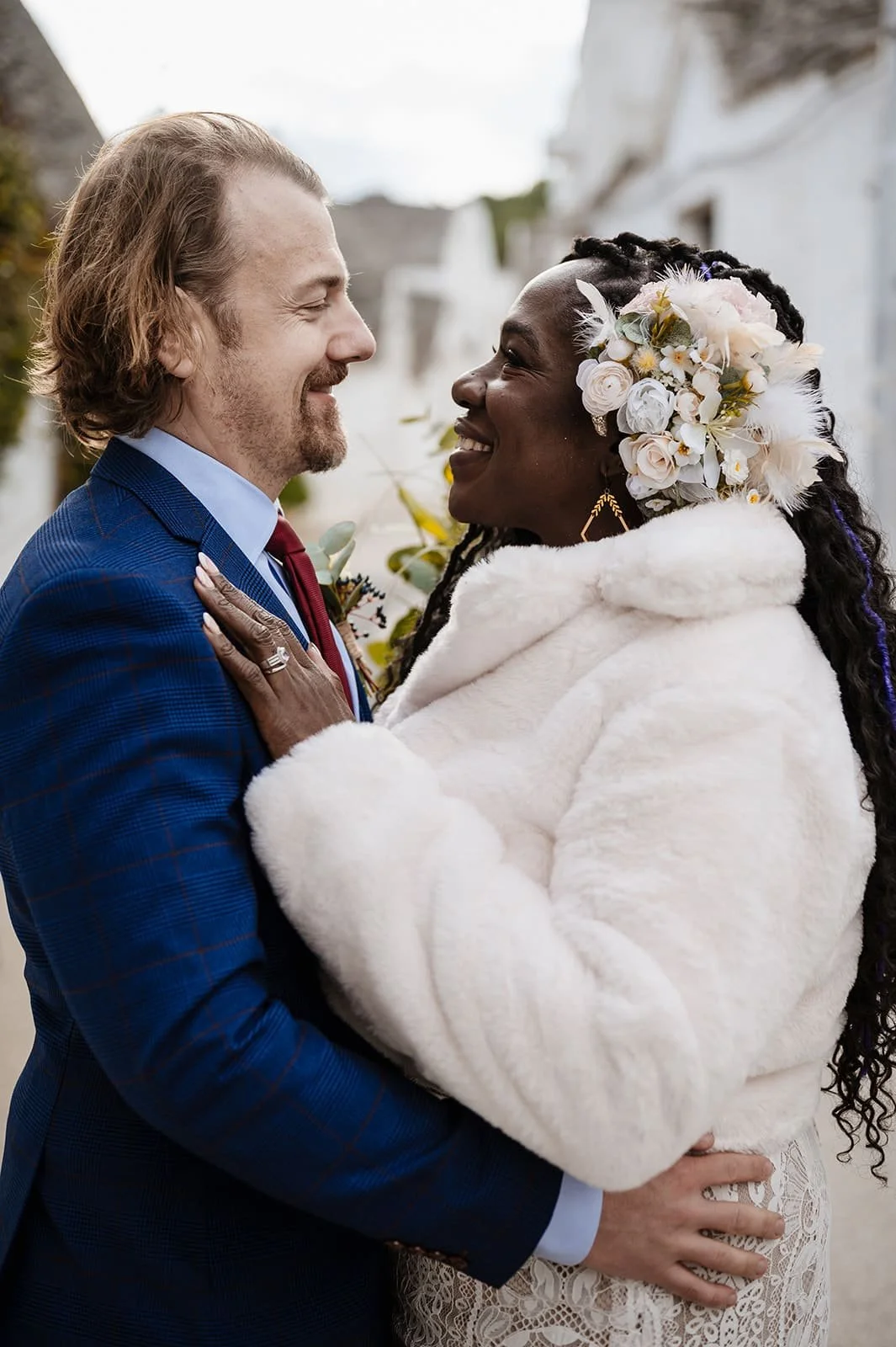 A couple on their wedding day, gazing into each other's eyes, with the woman wearing a floral headpiece and a white fur coat, and the man in a blue suit.
