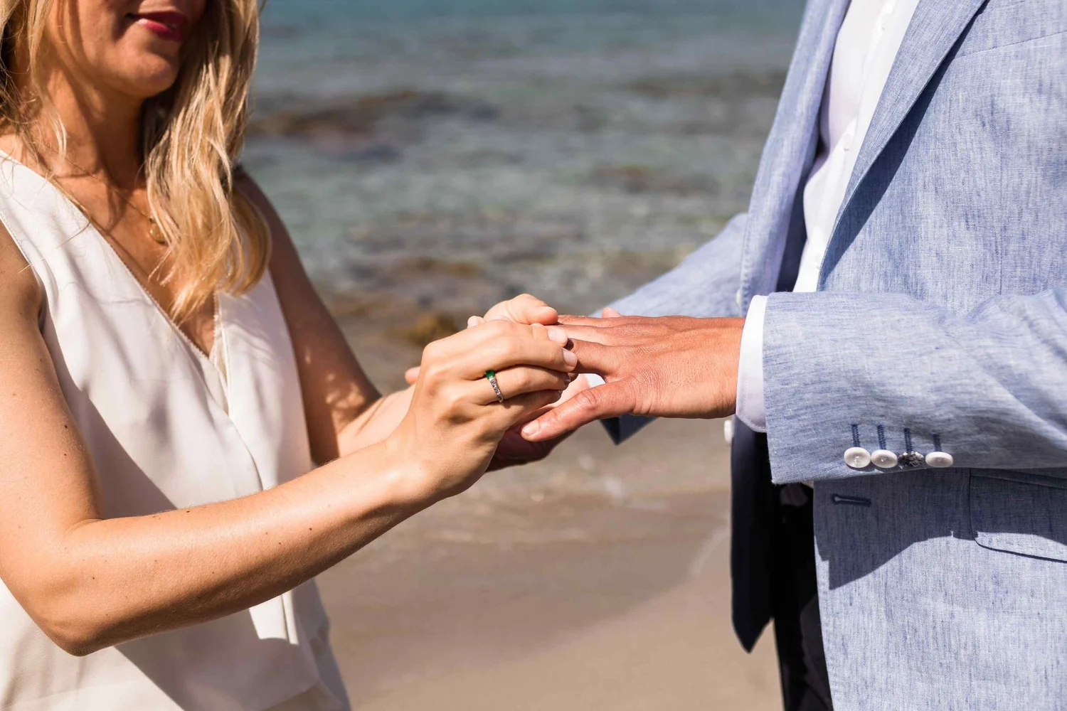 A woman and a man holding hands on the beach, with the woman wearing a white dress and the man wearing a light blue suit.
