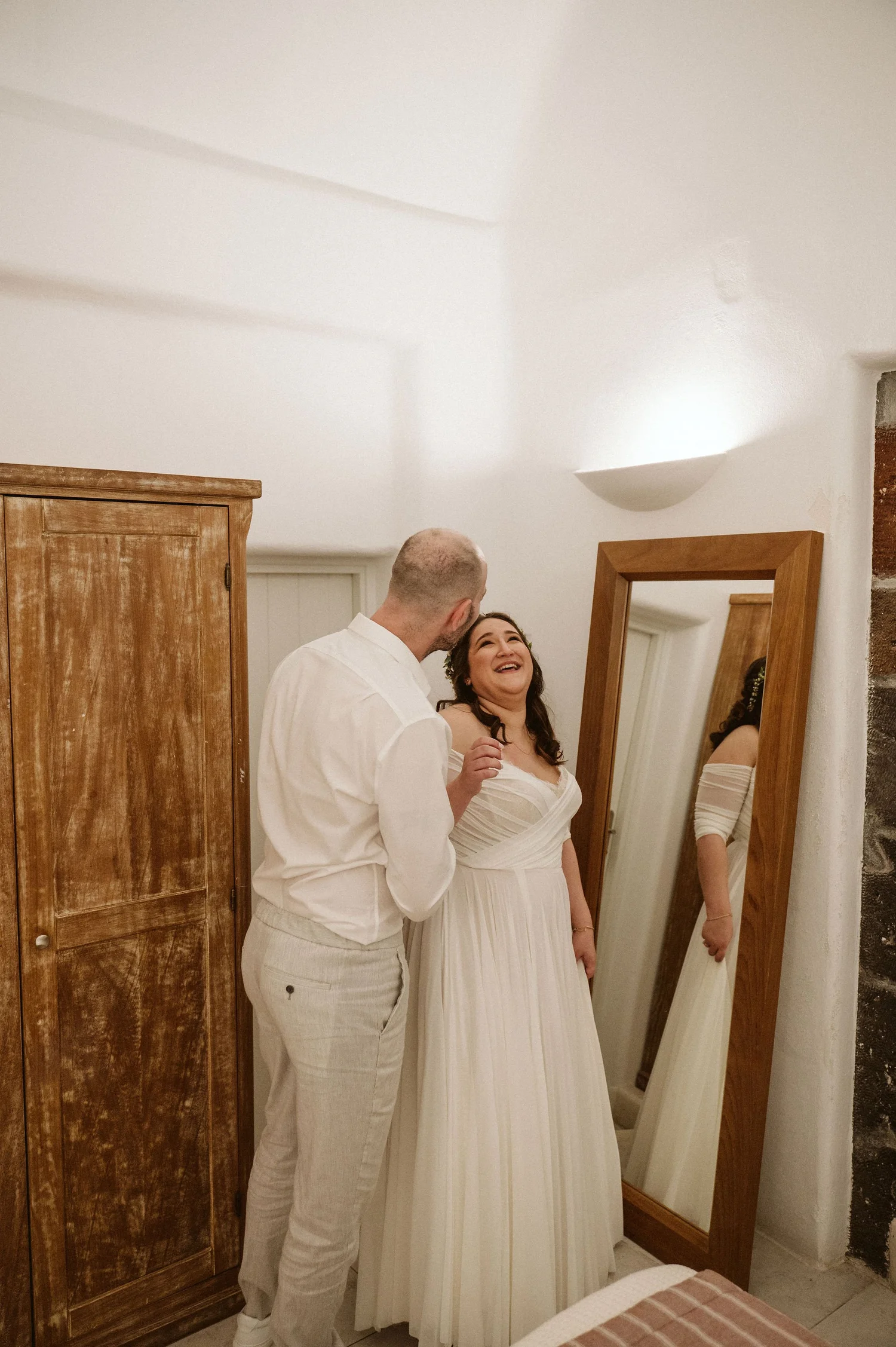 A man and woman smiling and looking at each other, standing in front of a mirror in a room with white walls. The woman is wearing a white dress, and the man is dressed in light-colored pants and a white shirt.