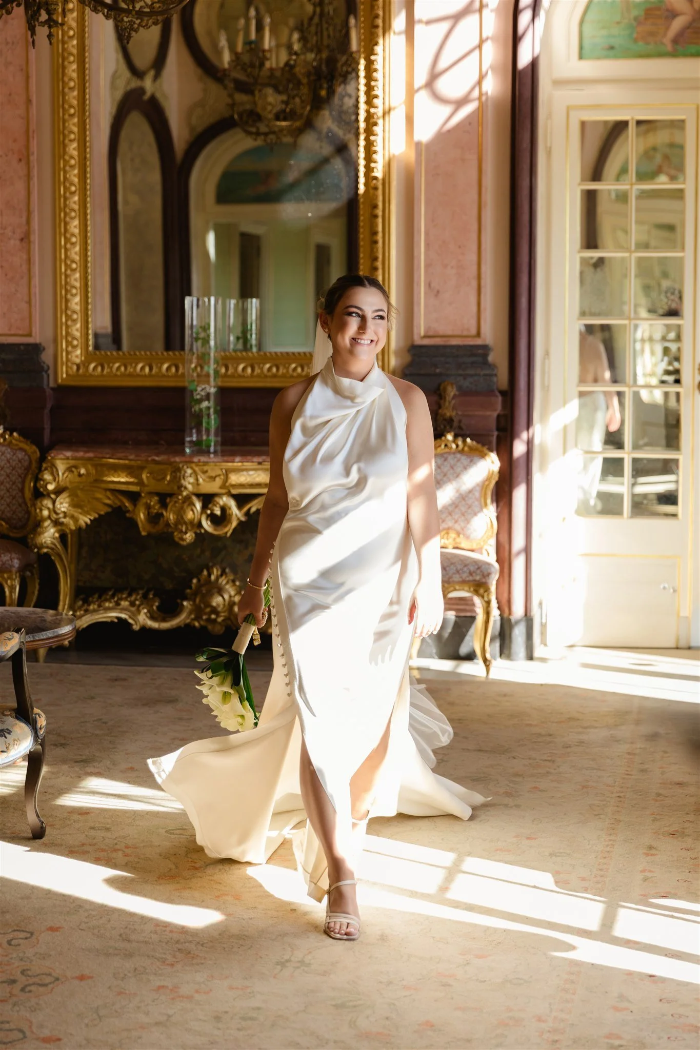 A woman in a white dress holding a small bouquet of white flowers standing in a luxurious room with gold and dark wood decor, large mirror, and sunlight streaming in through glass doors.