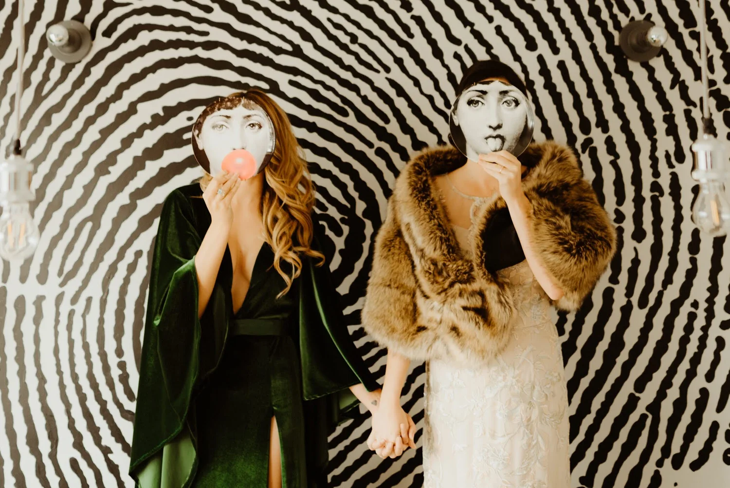 Two women in vintage fashion poses with mirrors covering their faces, standing in front of a bold, black-and-white zebra-striped background. One woman has long wavy hair and is holding a mirror with a pink bubblegum, while the other woman wears a fur