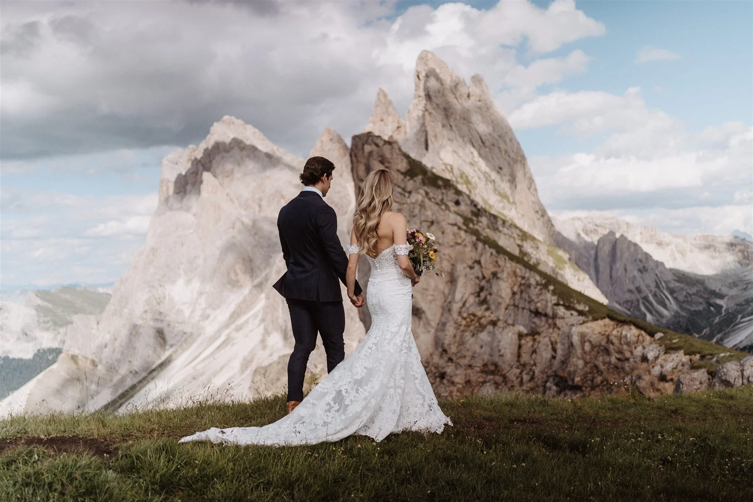A newlywed couple holding hands and walking in a mountainous outdoor setting, with rocky peaks and a partly cloudy sky in the background. The bride is wearing a white lace wedding gown and holding a bouquet, while the groom is in a dark suit.
