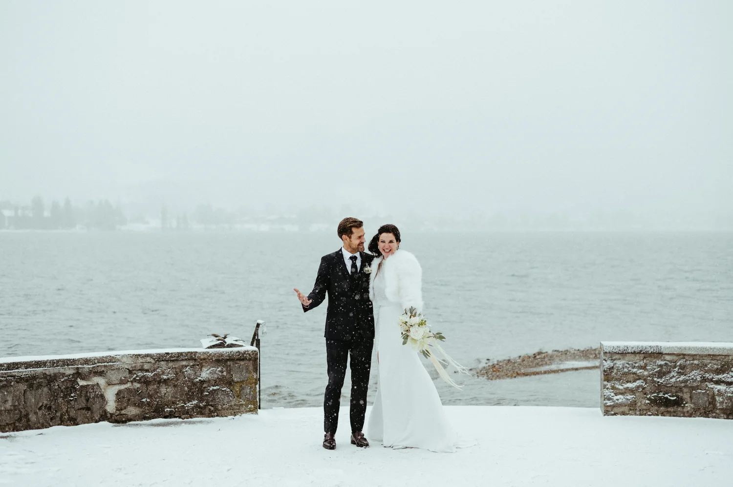 A bride and groom standing by a body of water on a snowy day, smiling and celebrating their wedding.