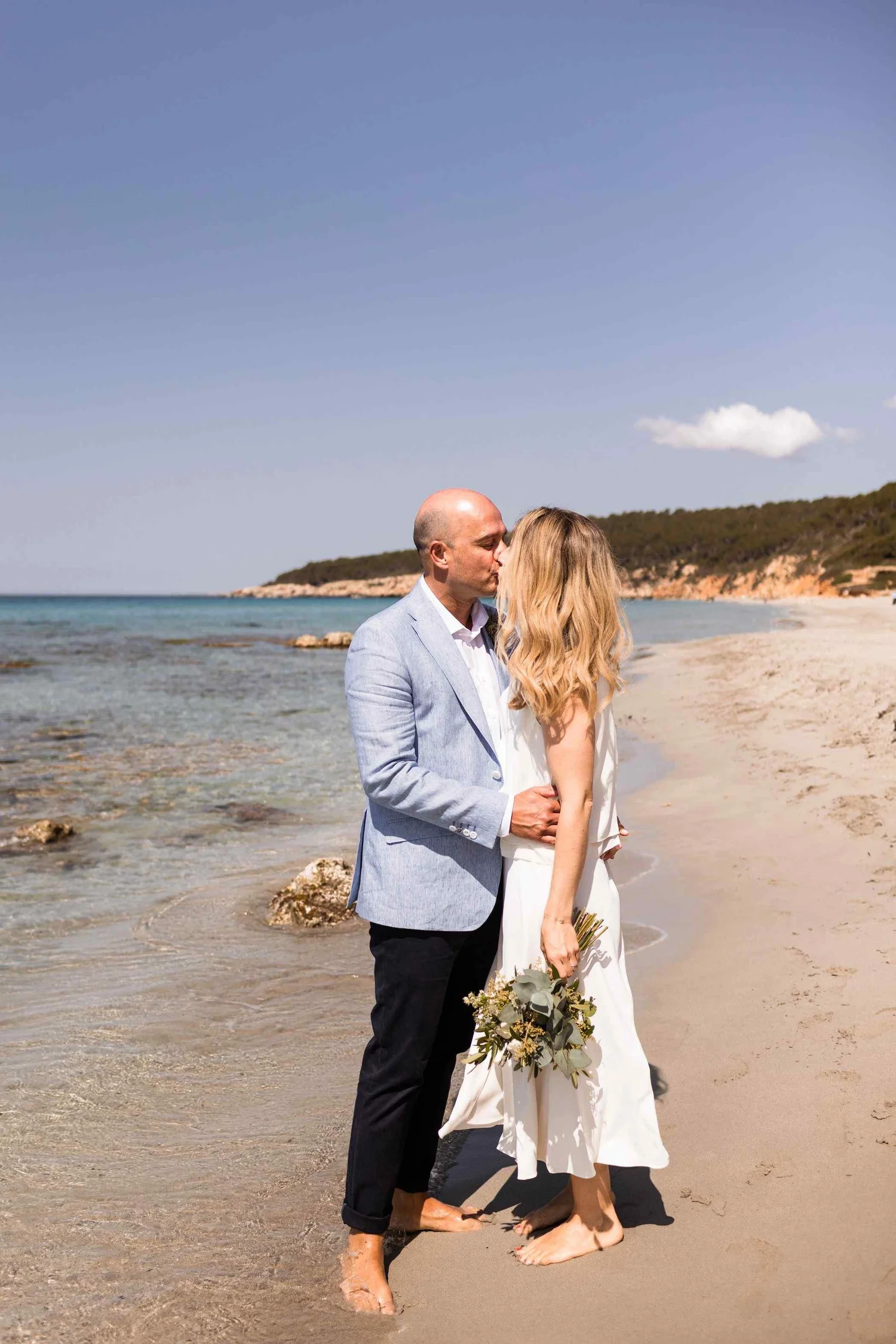 A couple stands barefoot on a sandy beach, kissing each other with the ocean and a distant shoreline with hills in the background. The man wears a light blue suit jacket and black pants, while the woman has long wavy blonde hair, a white dress, and h
