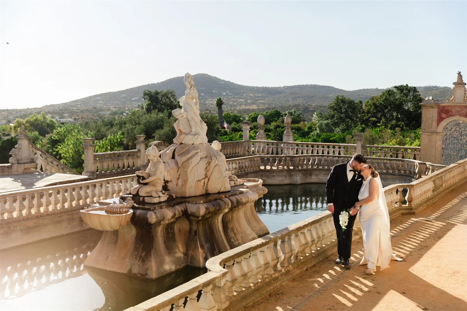 A bride and groom standing together on a terrace near a marble fountain with sculptures, overlooking a landscape with trees, hills, and clear sky.